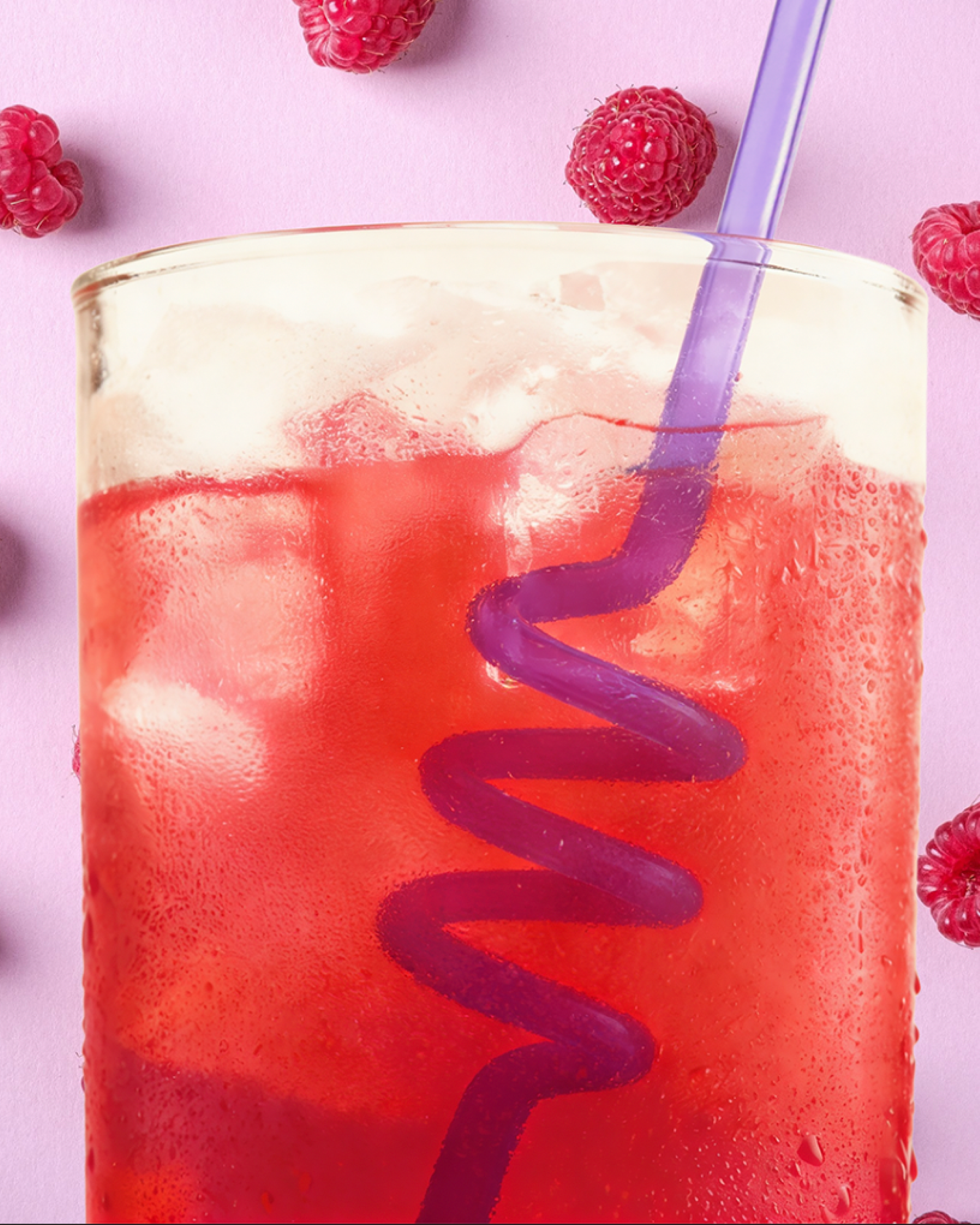 A close-up of a glass filled with pink raspberry-flavored beverage with ice, with a purple straw, on a pink background decorated with fresh raspberries.