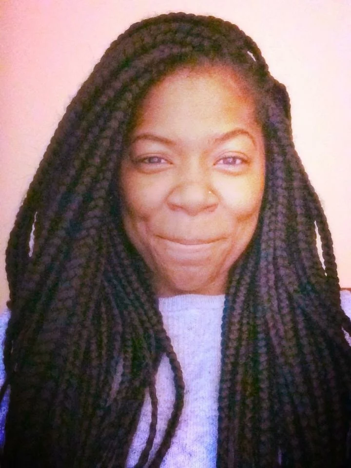 Brittinee, a cisgender neurodiverse Black woman, smiles at the camera with pursed lips while wearing a purple sweater