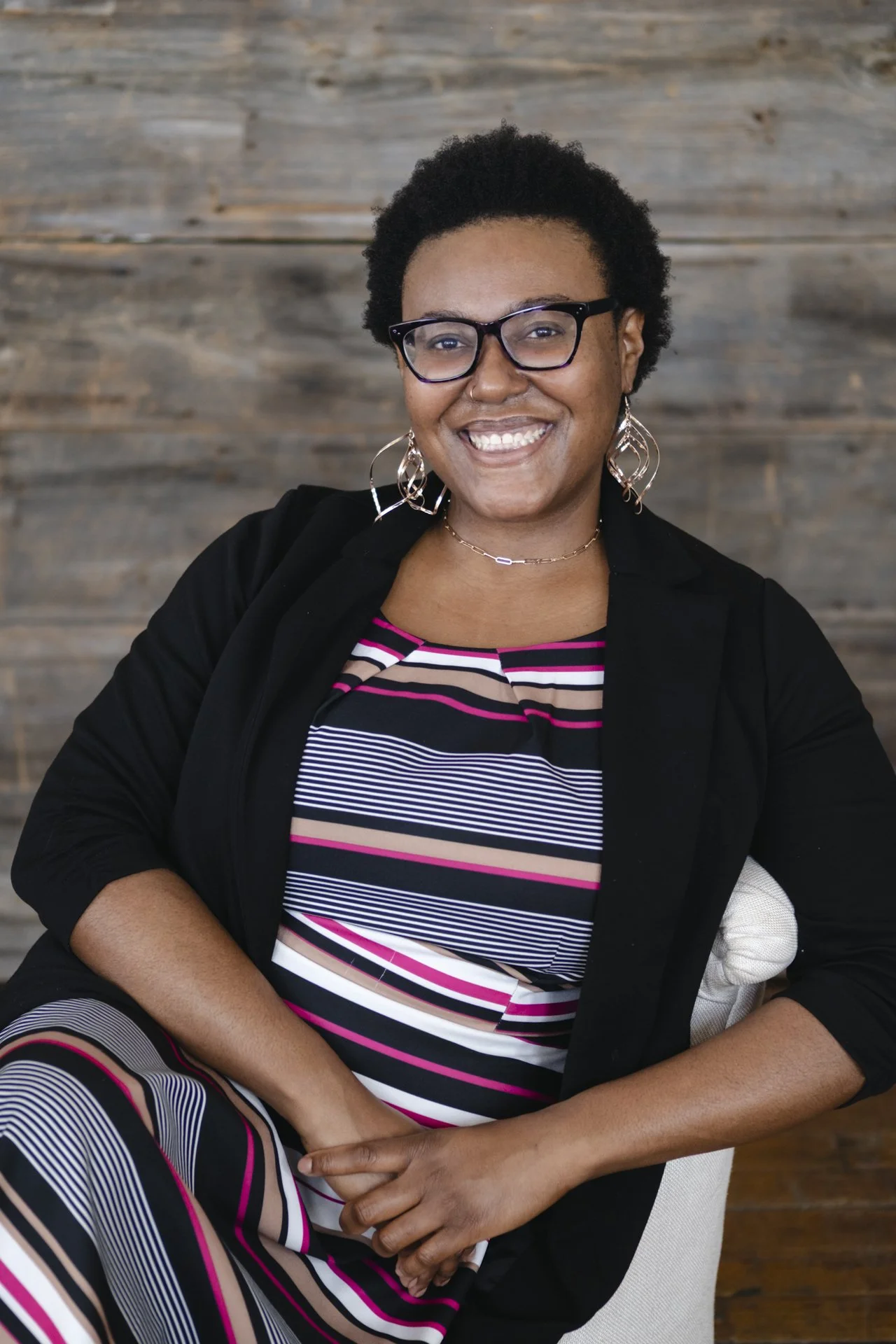 Smiling person with braided hair, wearing glasses and a light blue top, standing outdoors with greenery in the background.