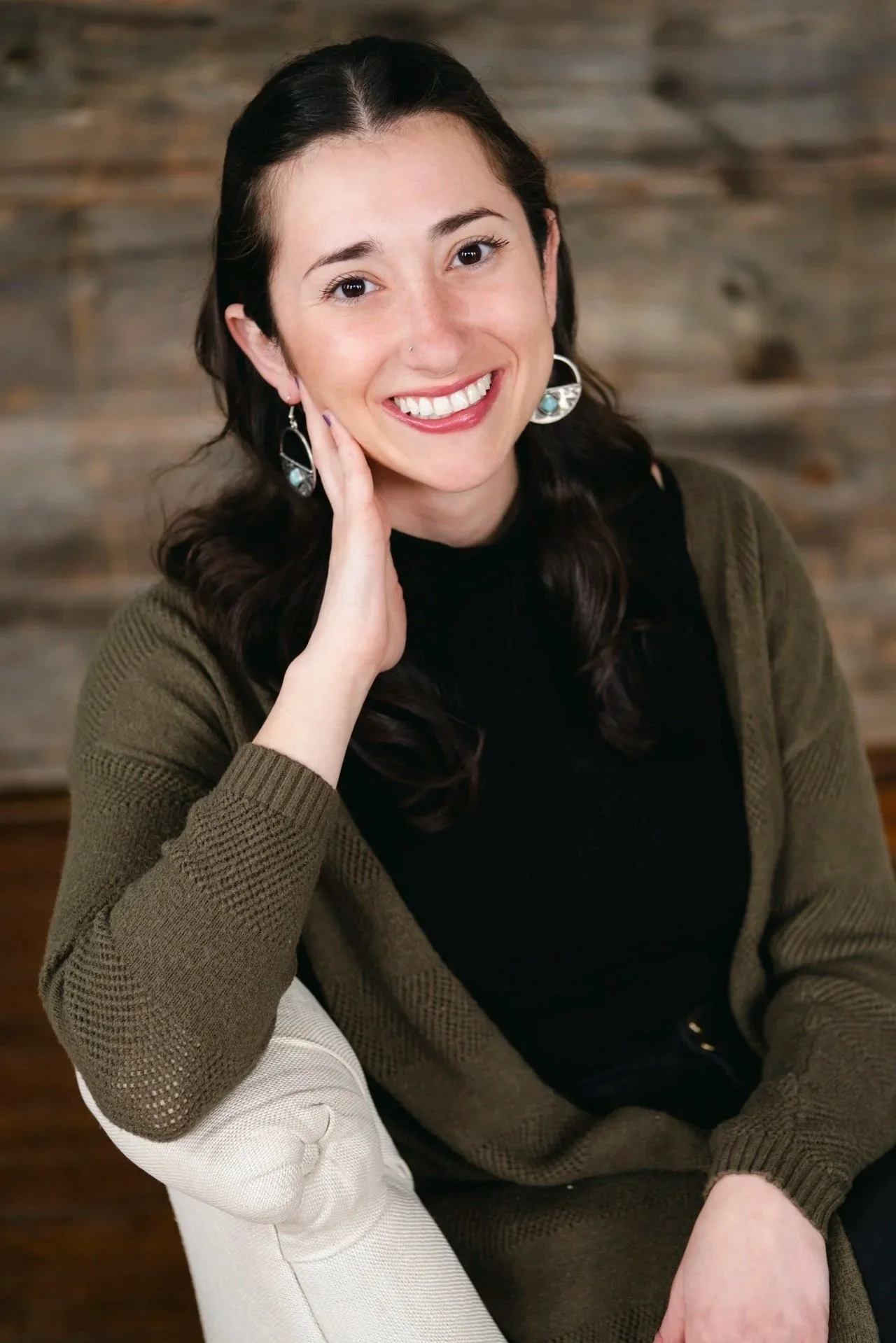 A woman with dark brown hair, smiling, wearing earrings, a black top, and an olive-green cardigan, sitting on a beige chair.