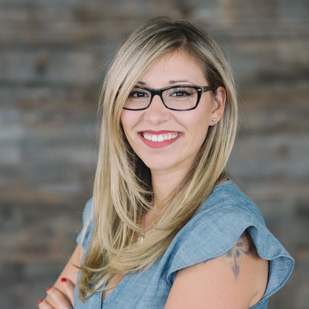 Woman with blonde hair and glasses smiling, wearing a blue top, with a tattoo on her arm, indoor setting.