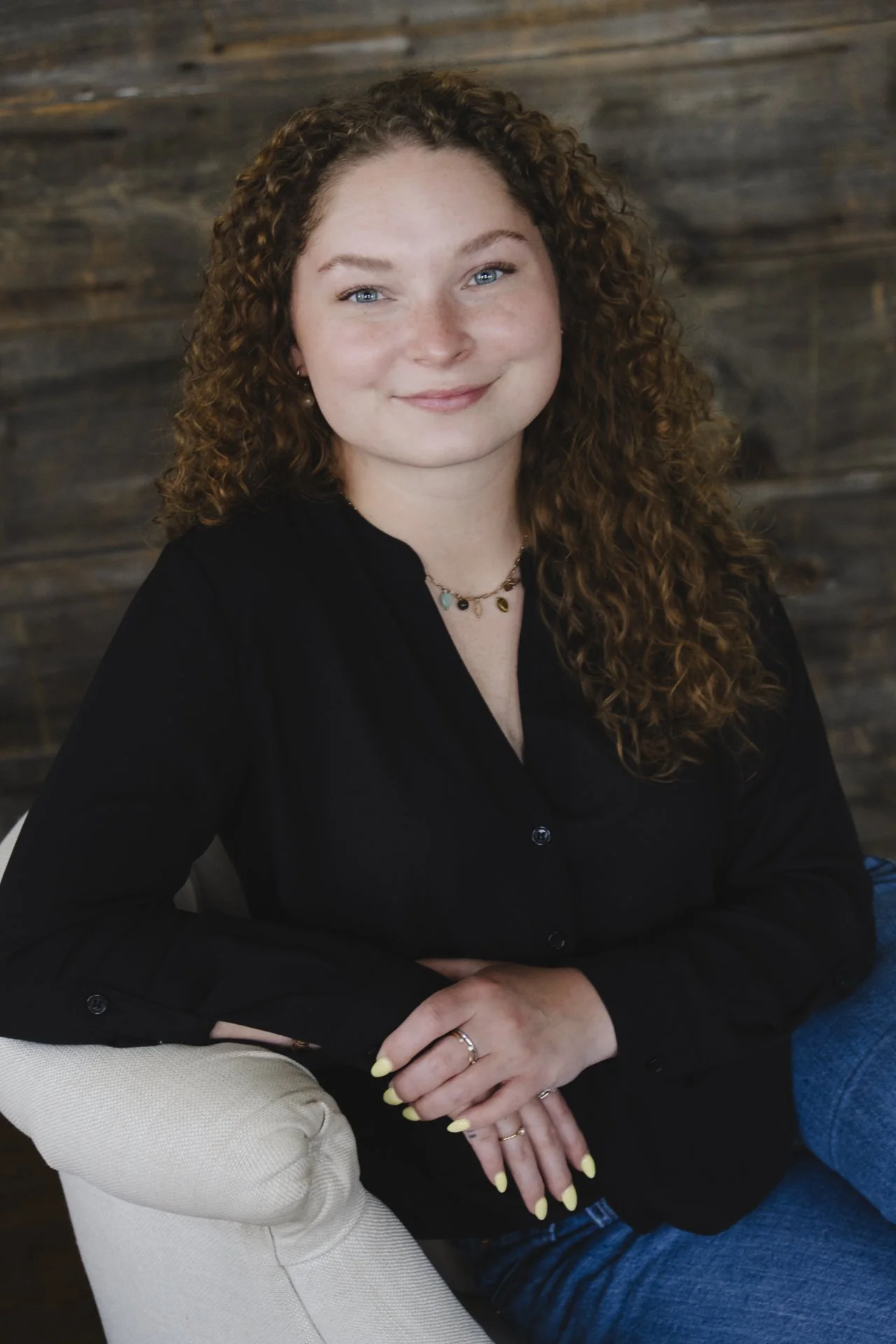 A young woman with curly red hair, blue eyes, and fair skin sitting on a white chair in front of a wooden wall. She is smiling and wearing a black blouse, a necklace, and rings on her fingers.