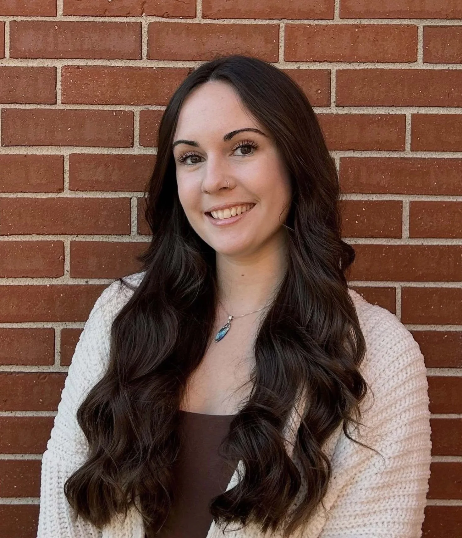 A young woman with long dark wavy hair, wearing a beige cardigan over a brown top, standing against a brick wall, smiling at the camera.