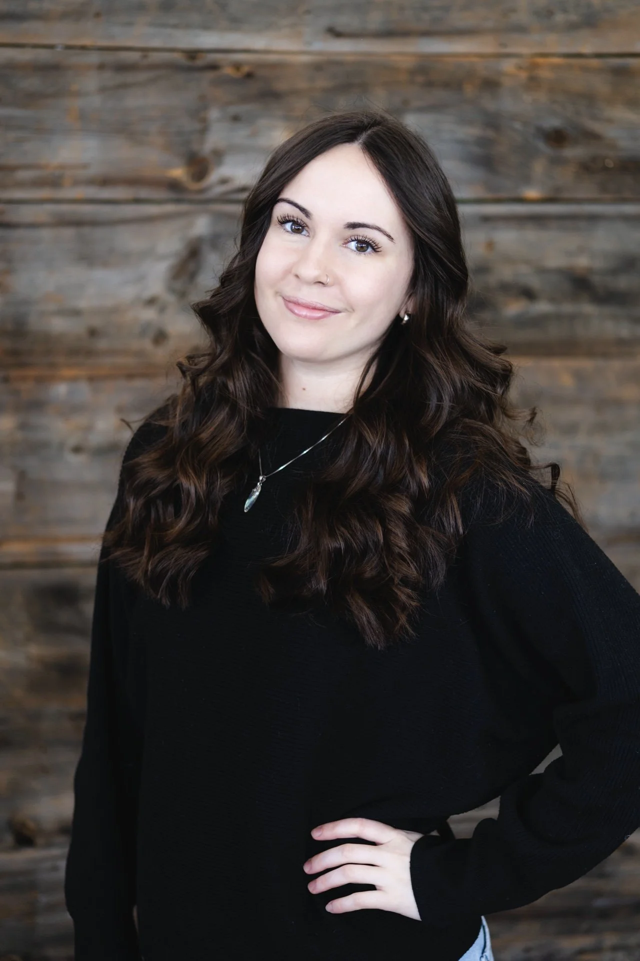 A young woman with long dark brown hair in loose curls, smiling, standing in front of a brick wall. She is wearing a beige knit cardigan over a dark top and a pendant necklace.