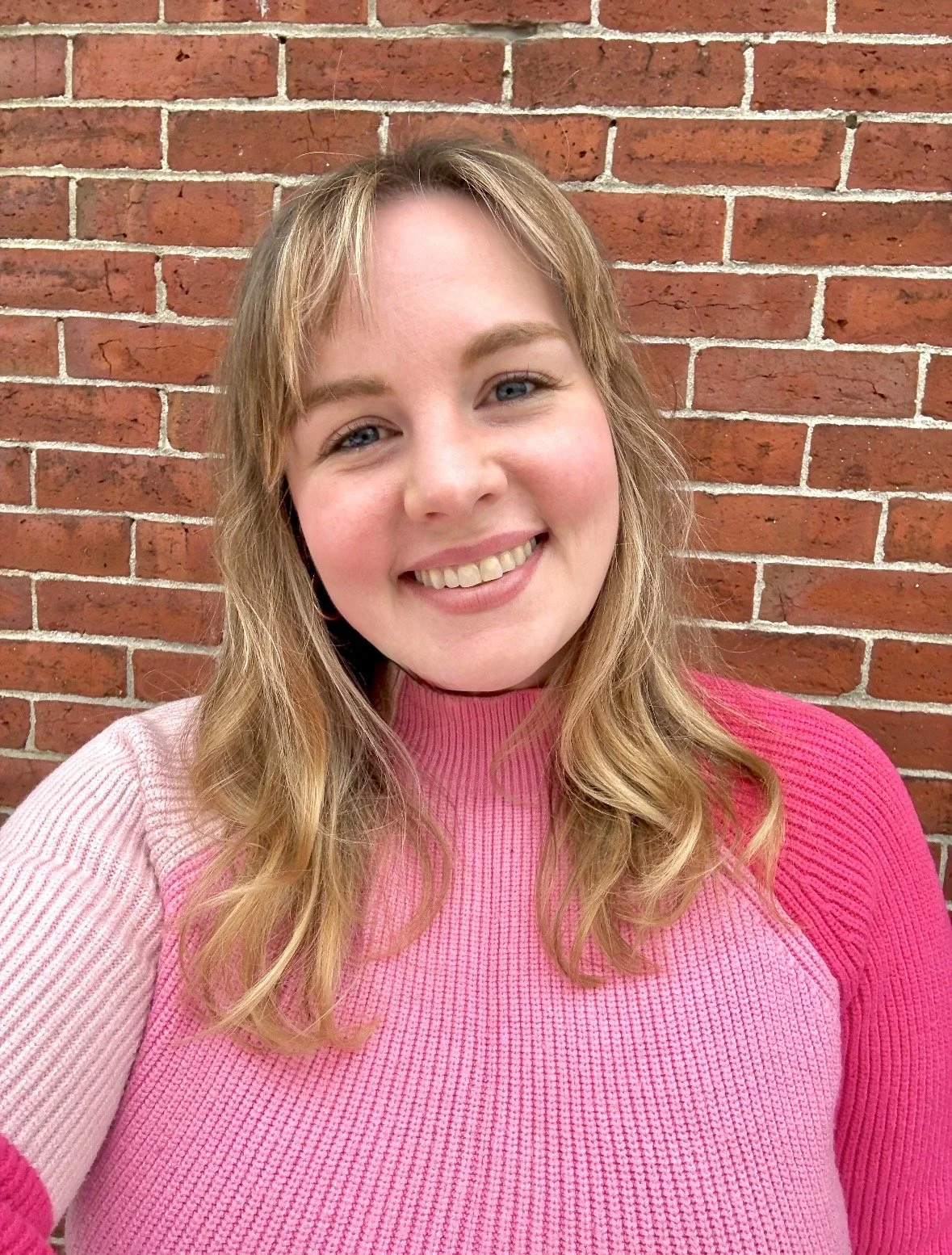 A young woman with blonde hair wearing a pink and beige sweater, smiling in front of a red brick wall.