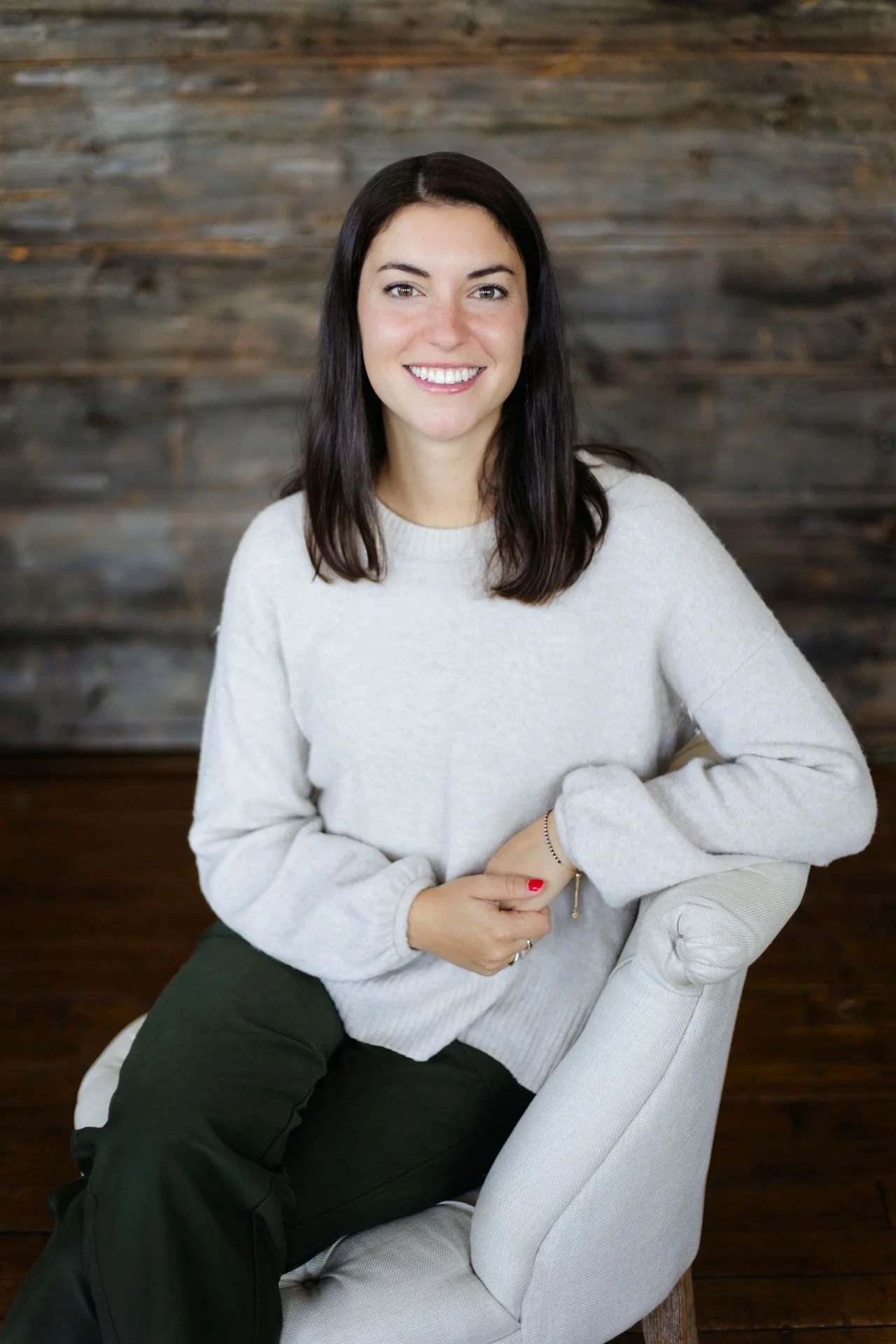 A woman with shoulder-length dark hair and a light gray sweater, smiling, sitting on a light-colored armchair in front of a wooden wall.