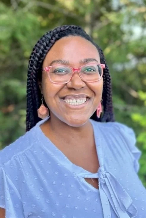 A woman with dark skin and braided hair smiling outdoors, wearing pink glasses and a light blue blouse with a bow detail, with a blurred green foliage background.