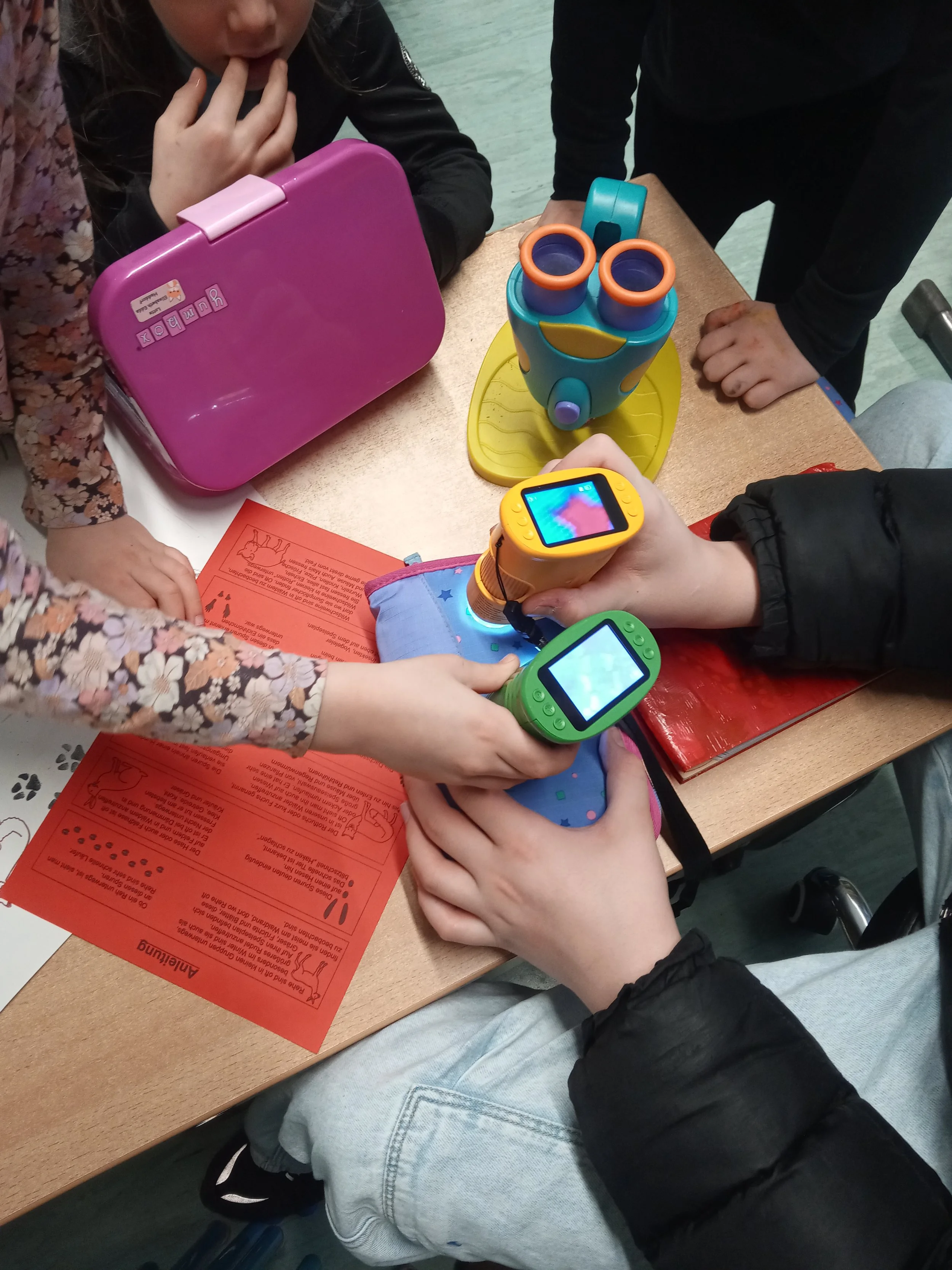 Kinder spielen mit Spielzeug und schauen auf einen analogen Thermometer auf einem Tisch, während sie eine rote Hinweisezettel und eine pinke Bücherbox haben.