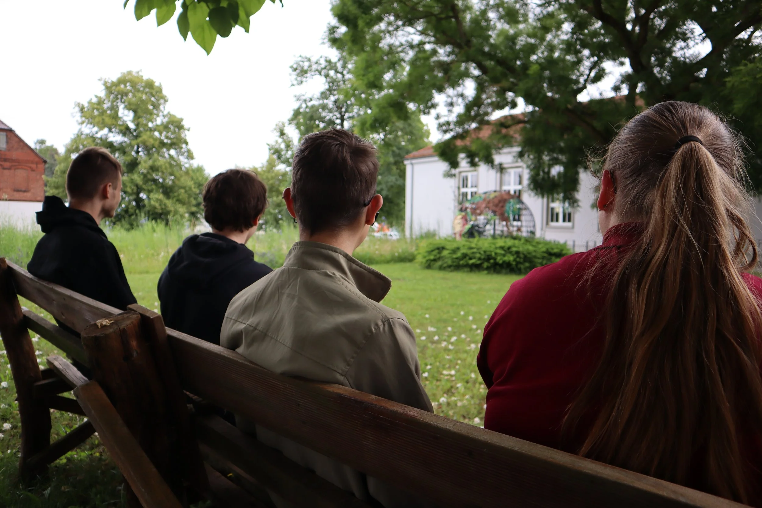 Vier Menschen sitzen auf einer Holzbank im Park, schauen in die Ferne, umgeben von grünen Bäumen, im Hintergrund ein weißes Haus.