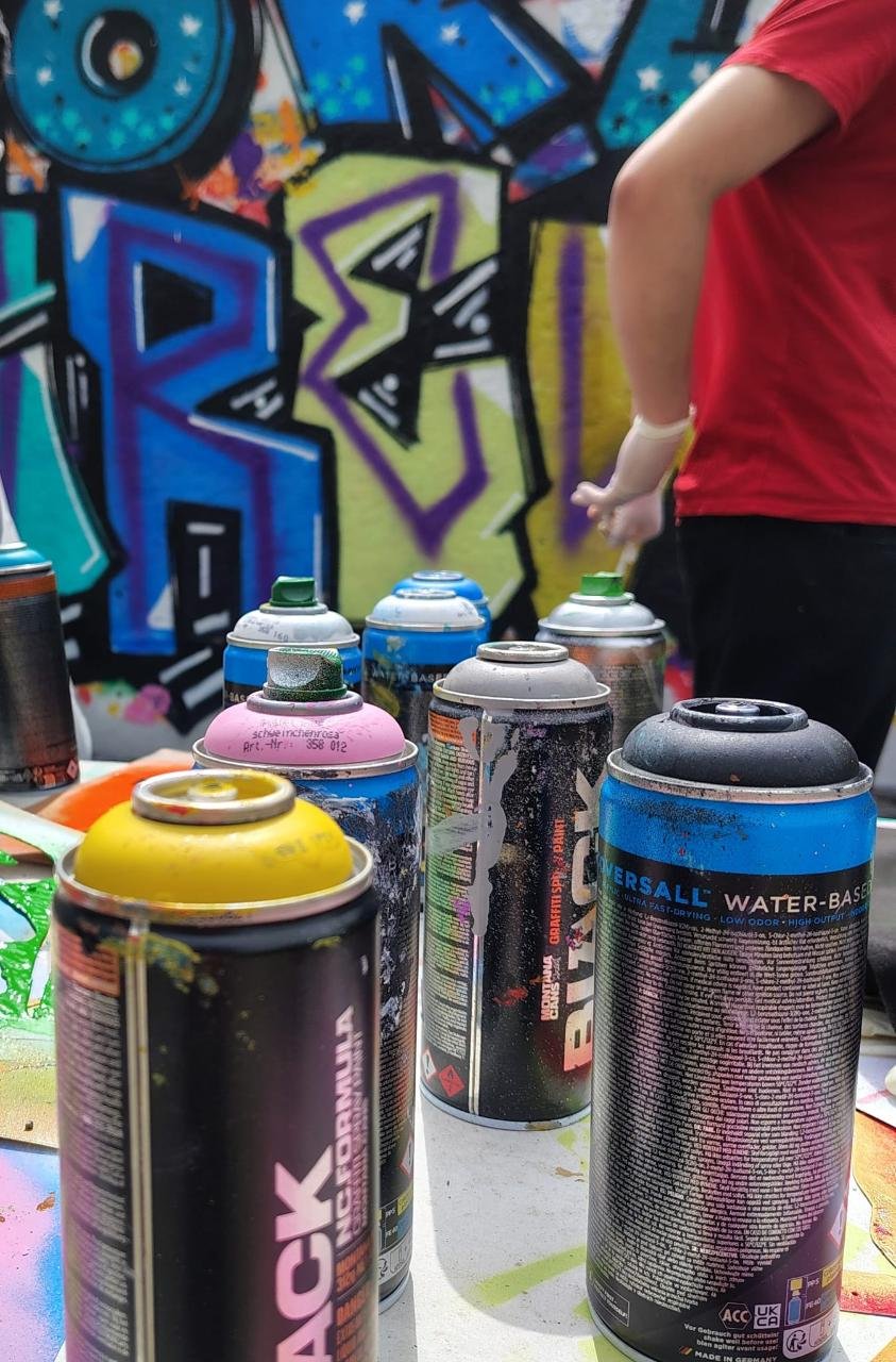 Mehrere Spraydosen stehen auf einem Tisch vor einer bunten Graffiti-Wand, eine Person trägt rote Kleidung und weiße Handschuhe.