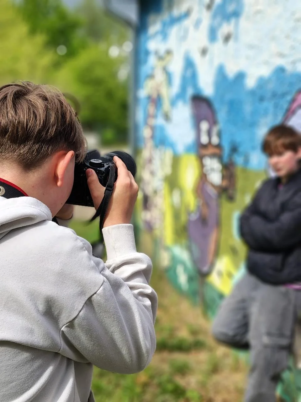 Ein Junge macht ein Foto von einem Mädchen vor einer bunten Wand mit Graffiti. Das Mädchen steht mit verschränkten Armen und trägt eine schwarze Jacke.