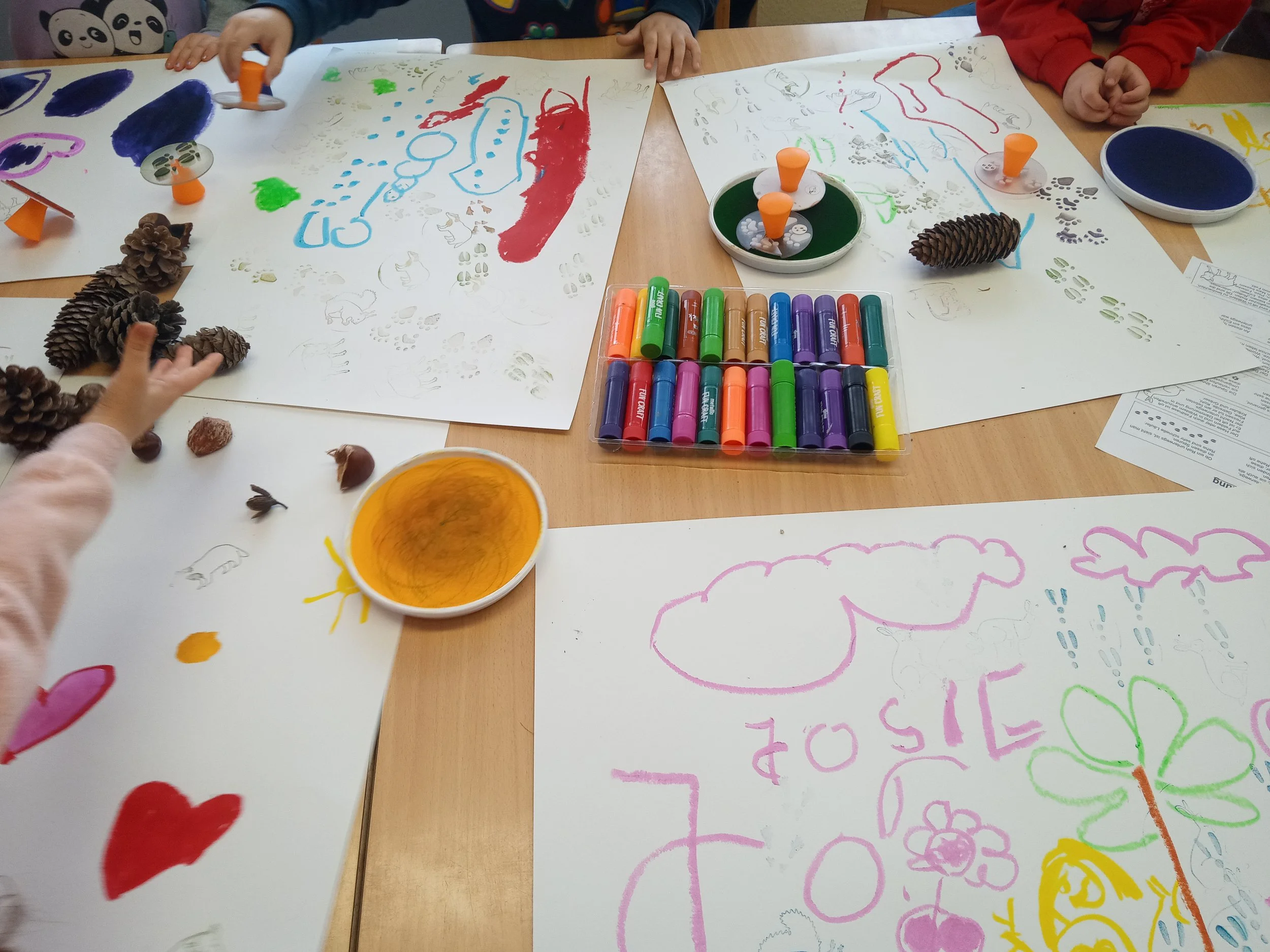Kinder beim gemeinsamen Malen und Basteln mit bunten Farben, Papier, Pinseln und Naturmaterialien auf einem Tisch.