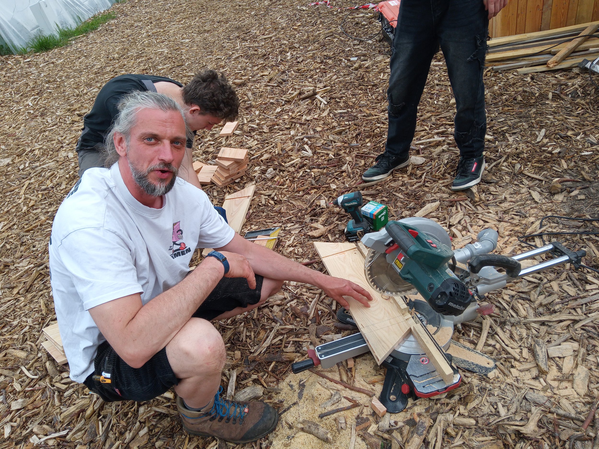 Drei Männer arbeiten im Garten mit Holz und Werkzeugen, überwiegend im Boden sitzend oder stehend, auf einer Fläche mit Holzspänen.