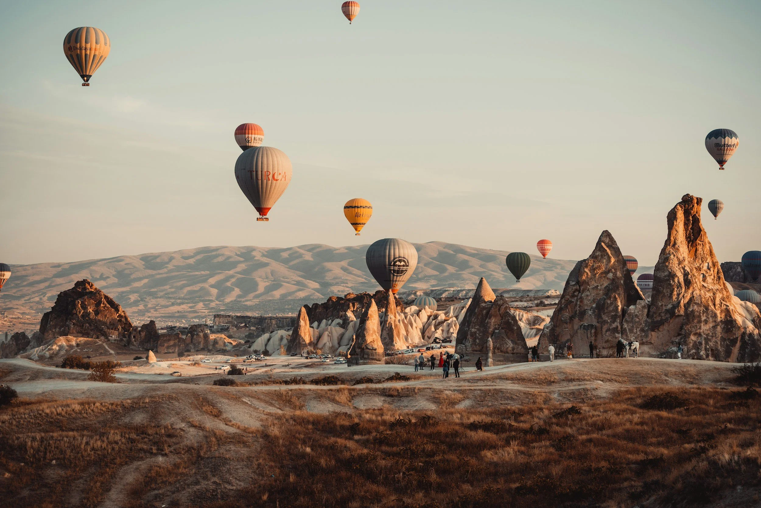 Multiple hot air balloons floating over a rocky landscape with unique rock formations and snow-capped hills in the background during sunset.
