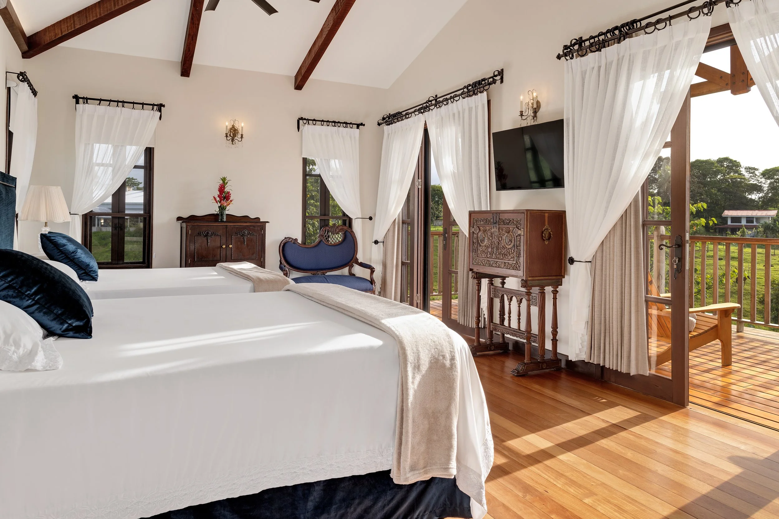 A bedroom with two beds, each with white bedspreads and blue pillows, wooden furniture, a TV mounted on the wall, large windows with white curtains, and a small balcony with wooden flooring outside.
