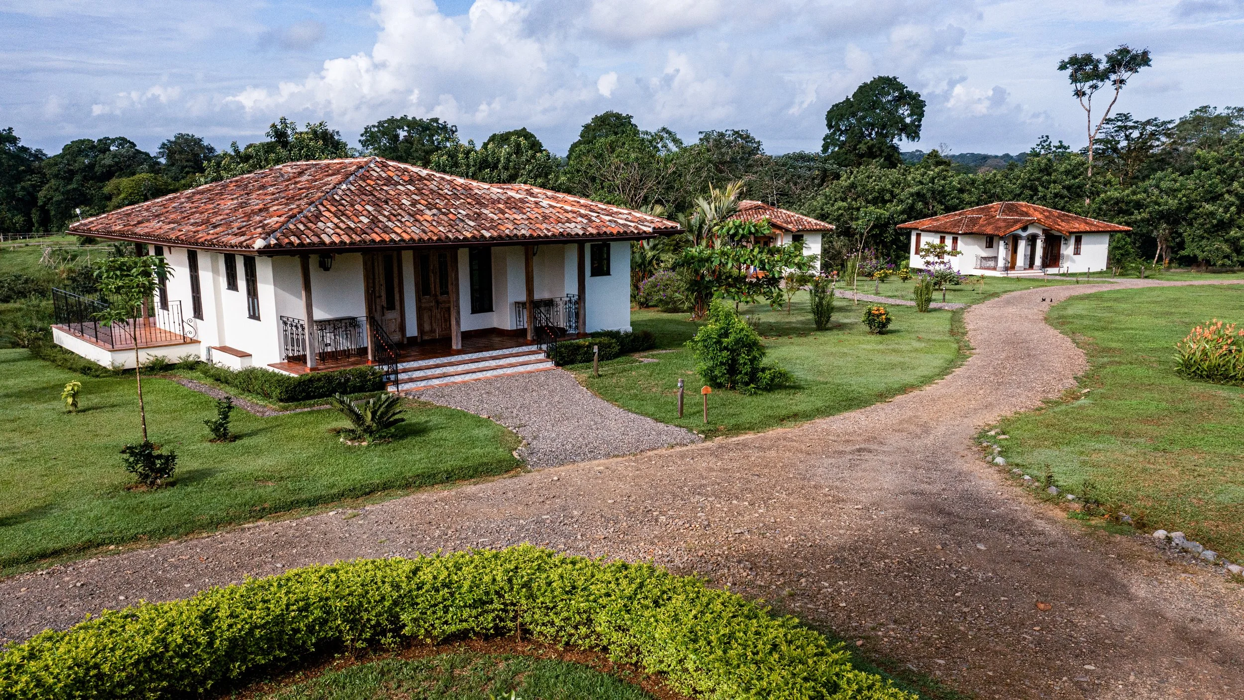 A scenic view of a white house with a red-tiled roof surrounded by green lawn, trees, and a gravel pathway leading towards other houses in the background.