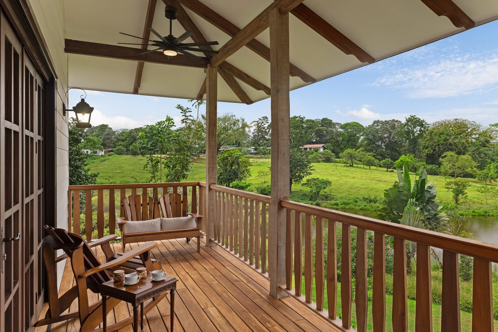 Balcony with wooden railing, outdoor furniture, and a ceiling fan overlooking lush green fields and trees under a partly cloudy sky.