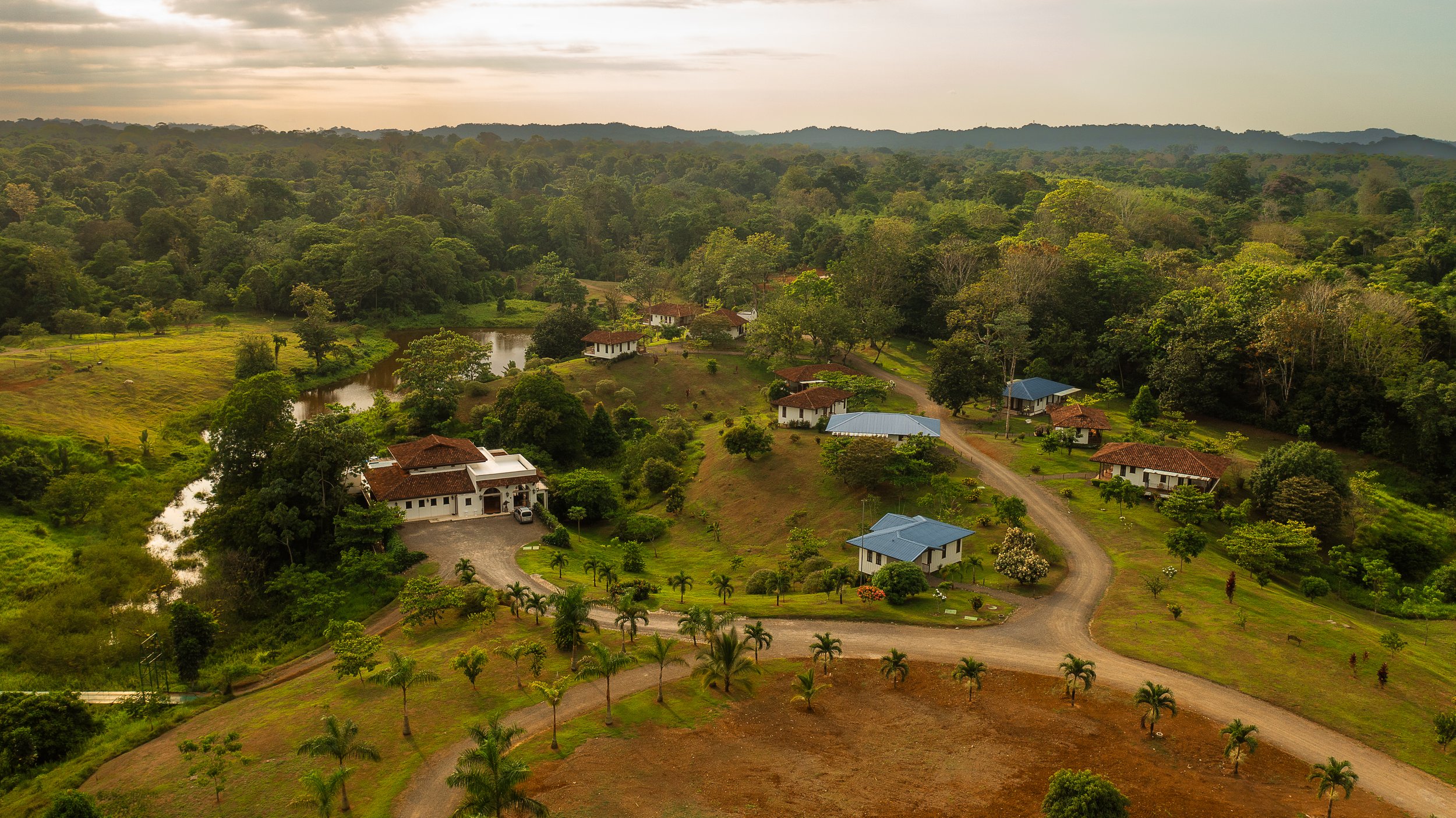 Aerial view of a lush green countryside with scattered houses, winding dirt roads, and a small pond, surrounded by dense forest and rolling hills under a cloudy sky.