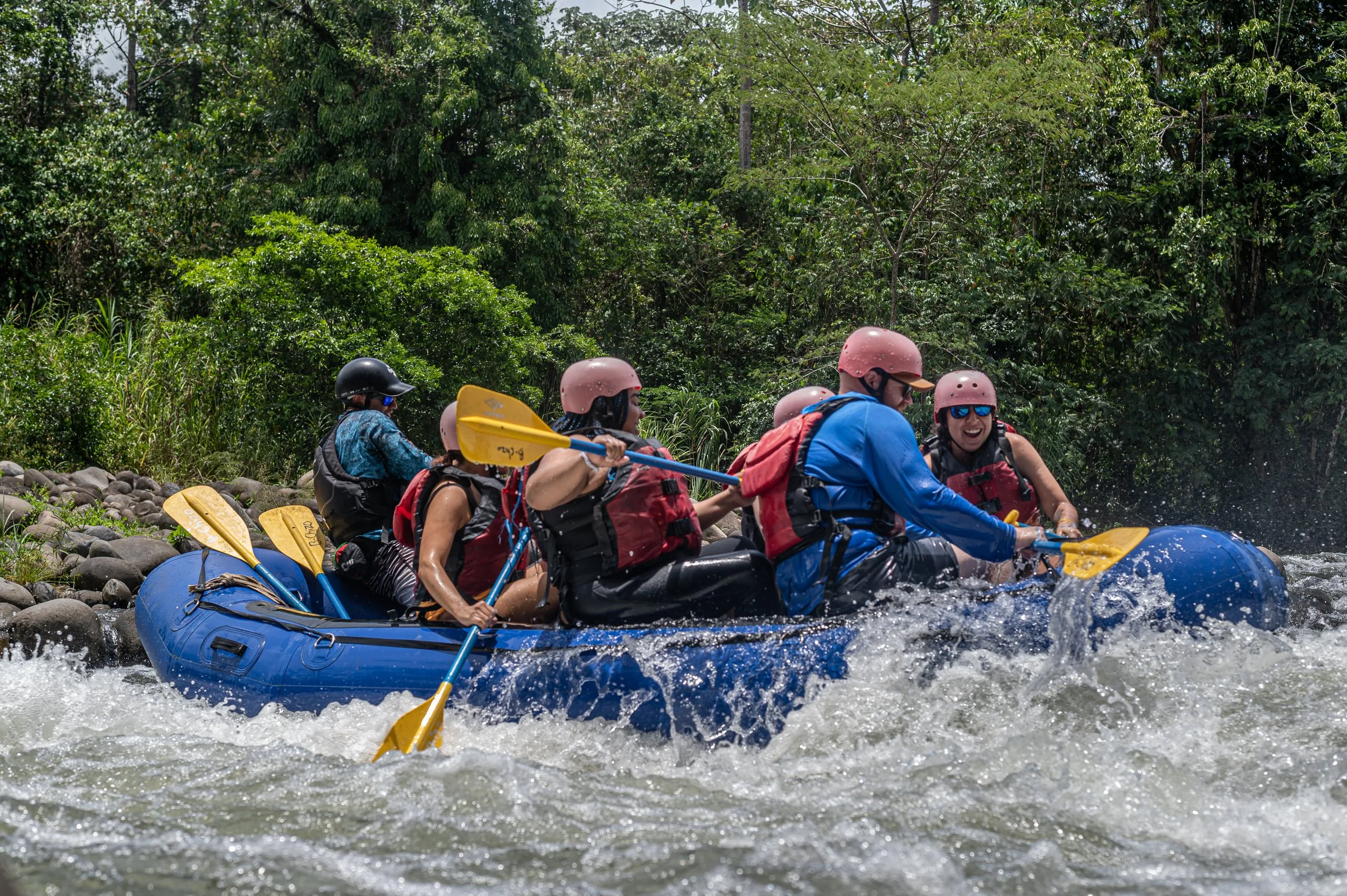 People are white water rafting on a river, wearing helmets and life jackets, with green trees in the background.