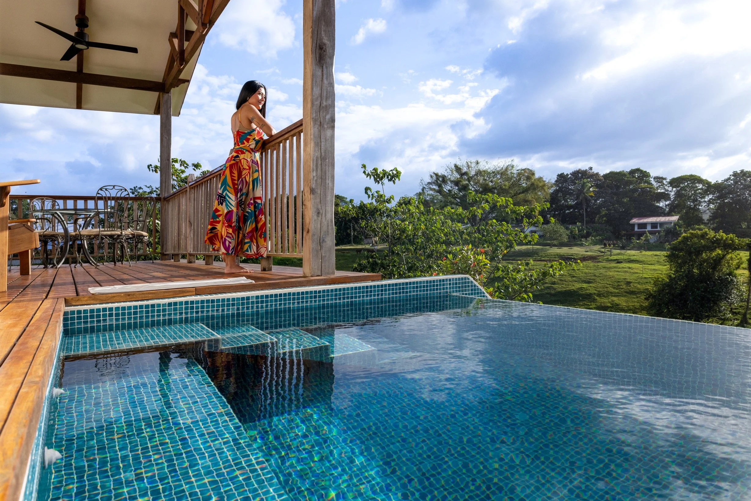 Woman standing on a wooden deck by a swimming pool, looking at scenic countryside with trees and a house in the distance, under partly cloudy sky.