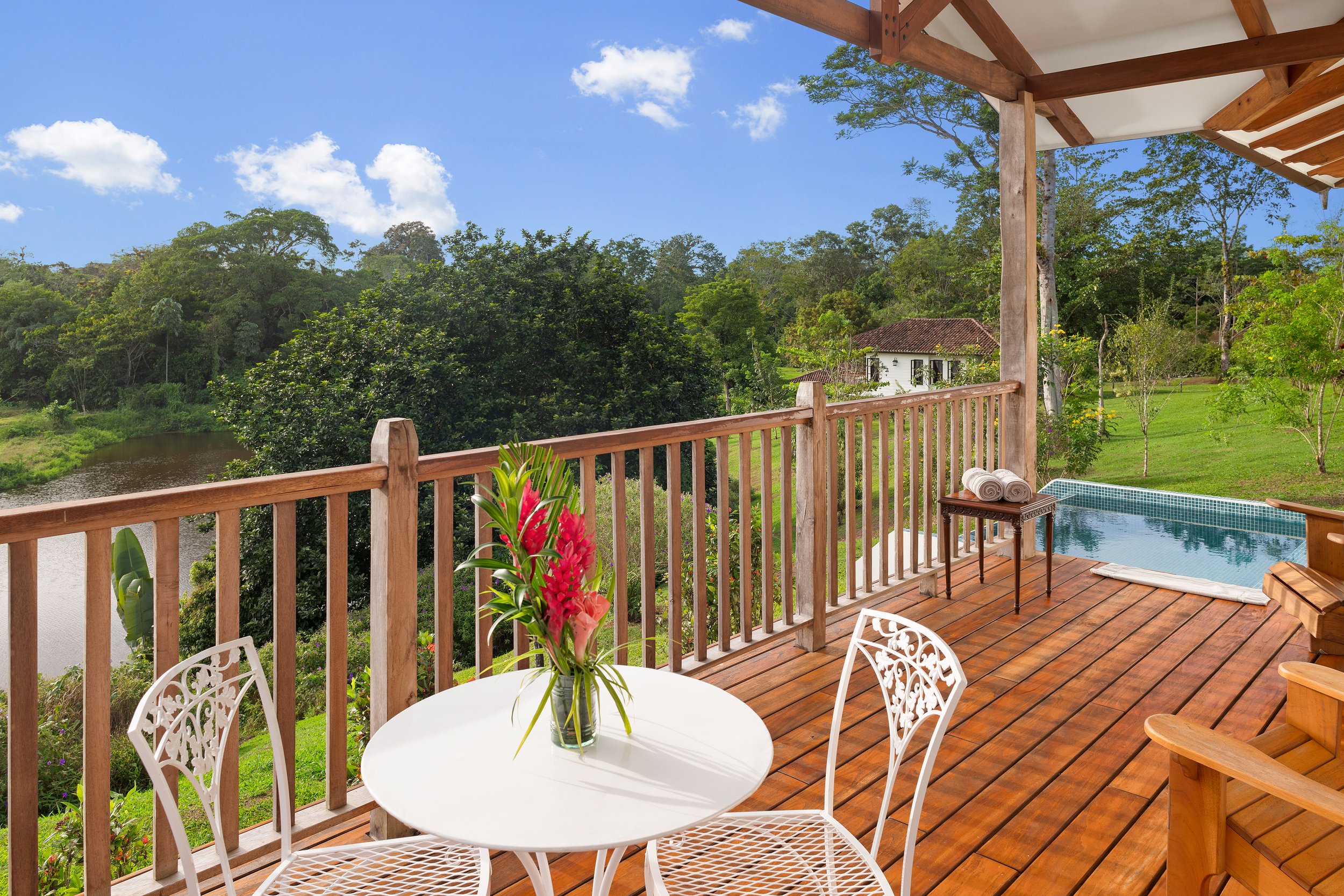 View of a wooden deck with a swimming pool, surrounded by green trees and a white house with a red tile roof in the distance. There is a round white table with floral arrangement, topped with towels, and white chairs around it.