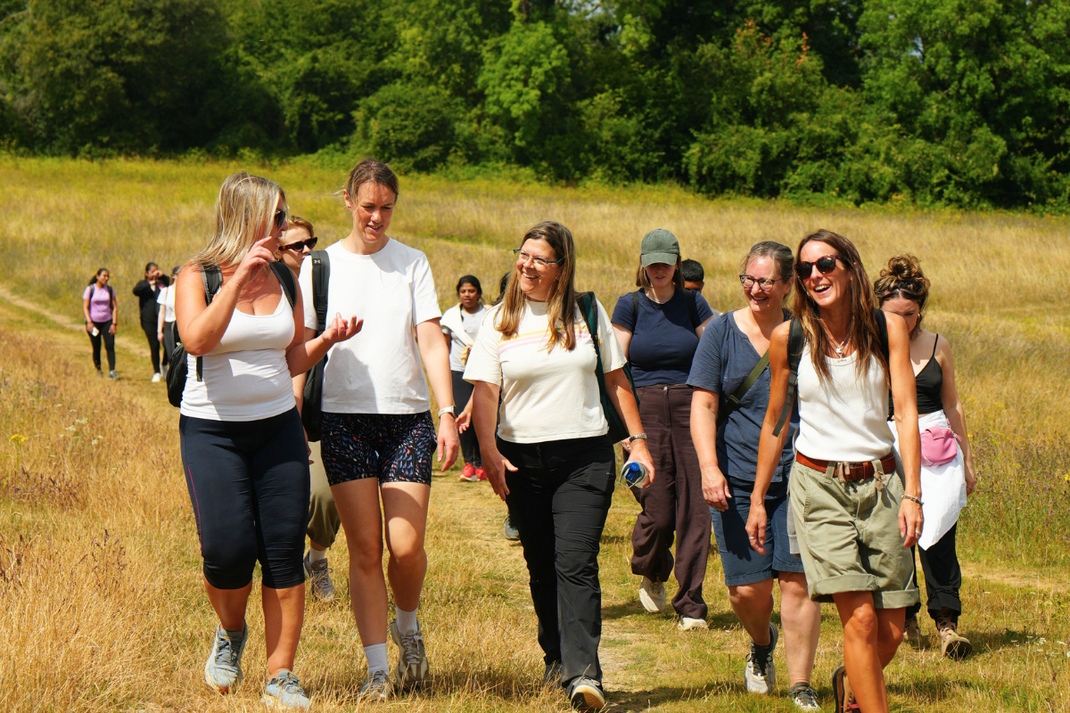 A group of women hiking outdoors in a grassy field with trees in the background, enjoying conversation and sunshine.