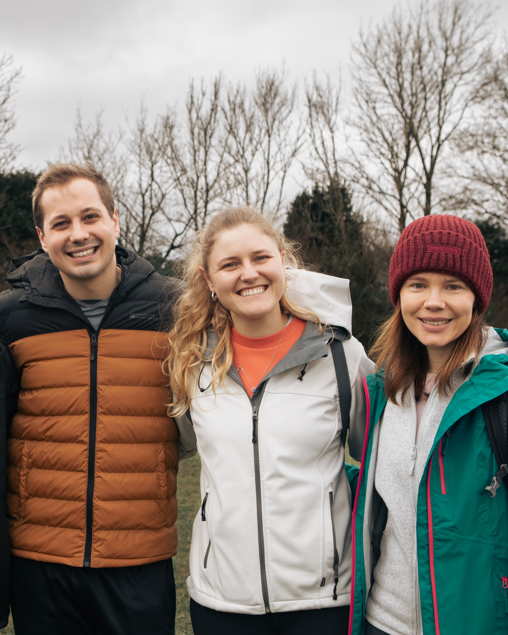 Three people standing outdoors in winter clothing with bare trees in the background, smiling at the camera.