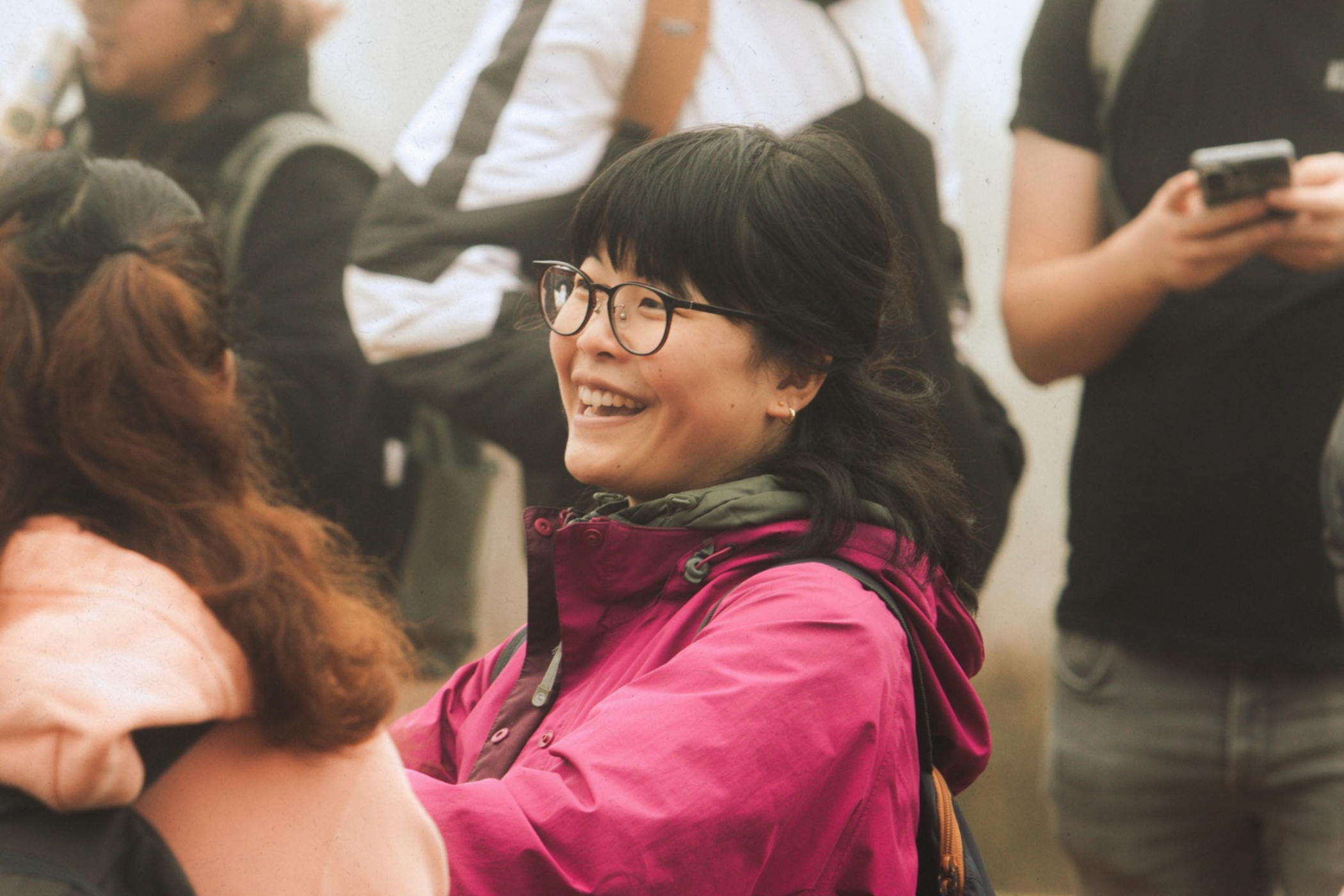 A woman with short dark hair, glasses, and earrings, smiling and talking to another person with long hair and a black sleeveless top in a crowd.