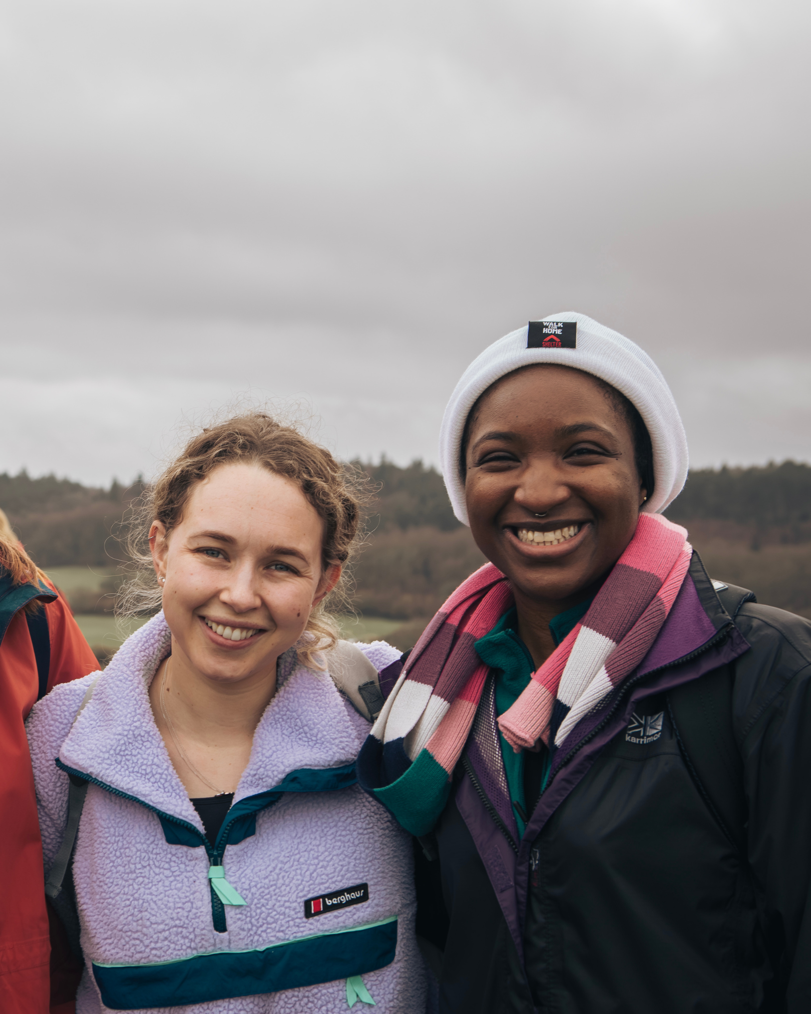 Close-up of two women smiling outdoors on a cloudy day, one wearing a purple fleece jacket and the other wearing a beanie and a striped scarf.