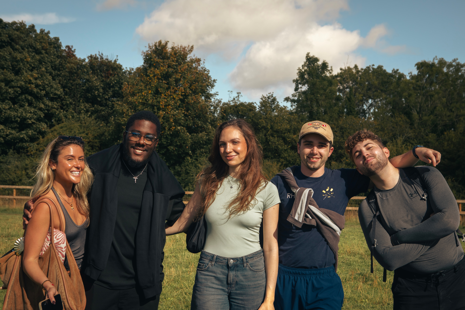 Group of five young adults standing outdoors in a park, smiling, with trees and a partly cloudy sky in the background.