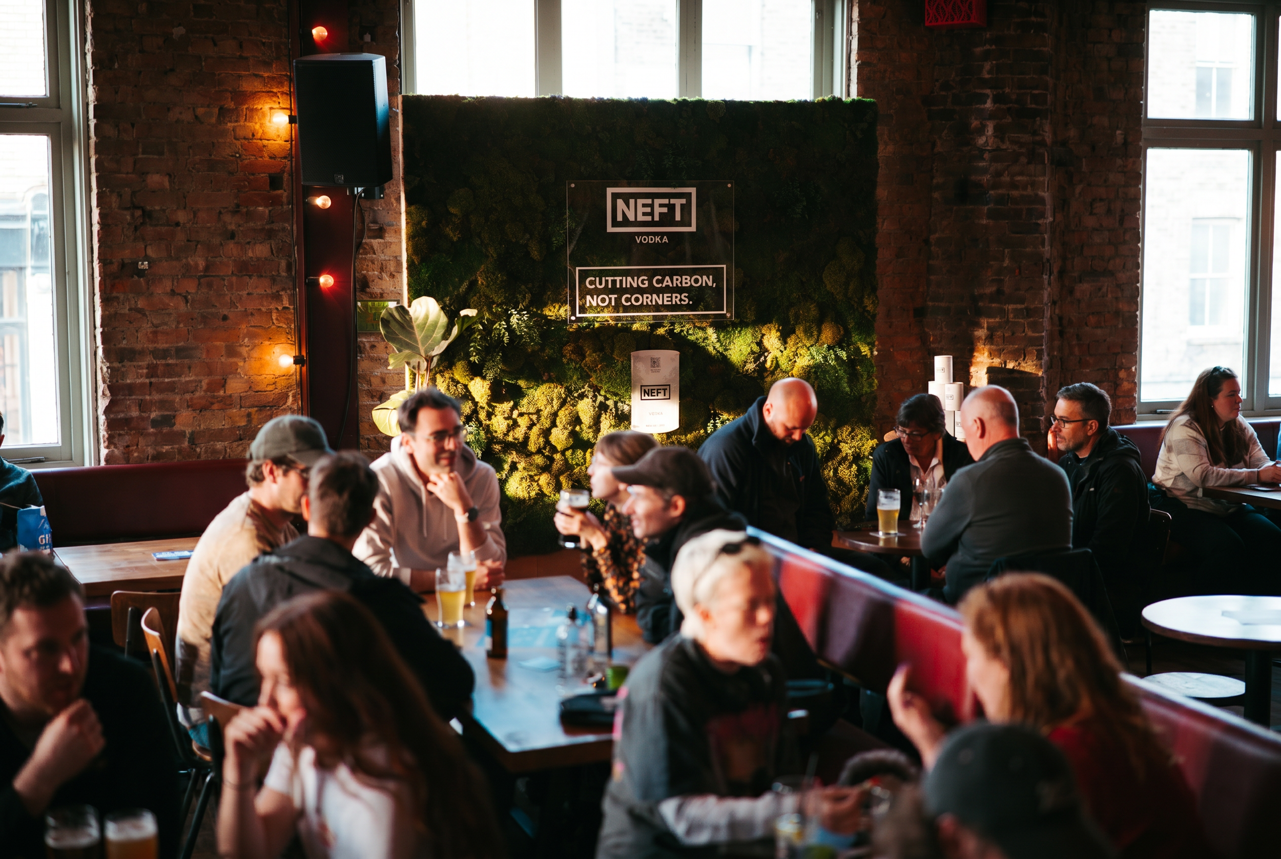 A group of people seated around tables inside a brick-walled venue, with a moss wall display advertising NEFT Vodka and the slogan 'Cutting carbon, not corners'.