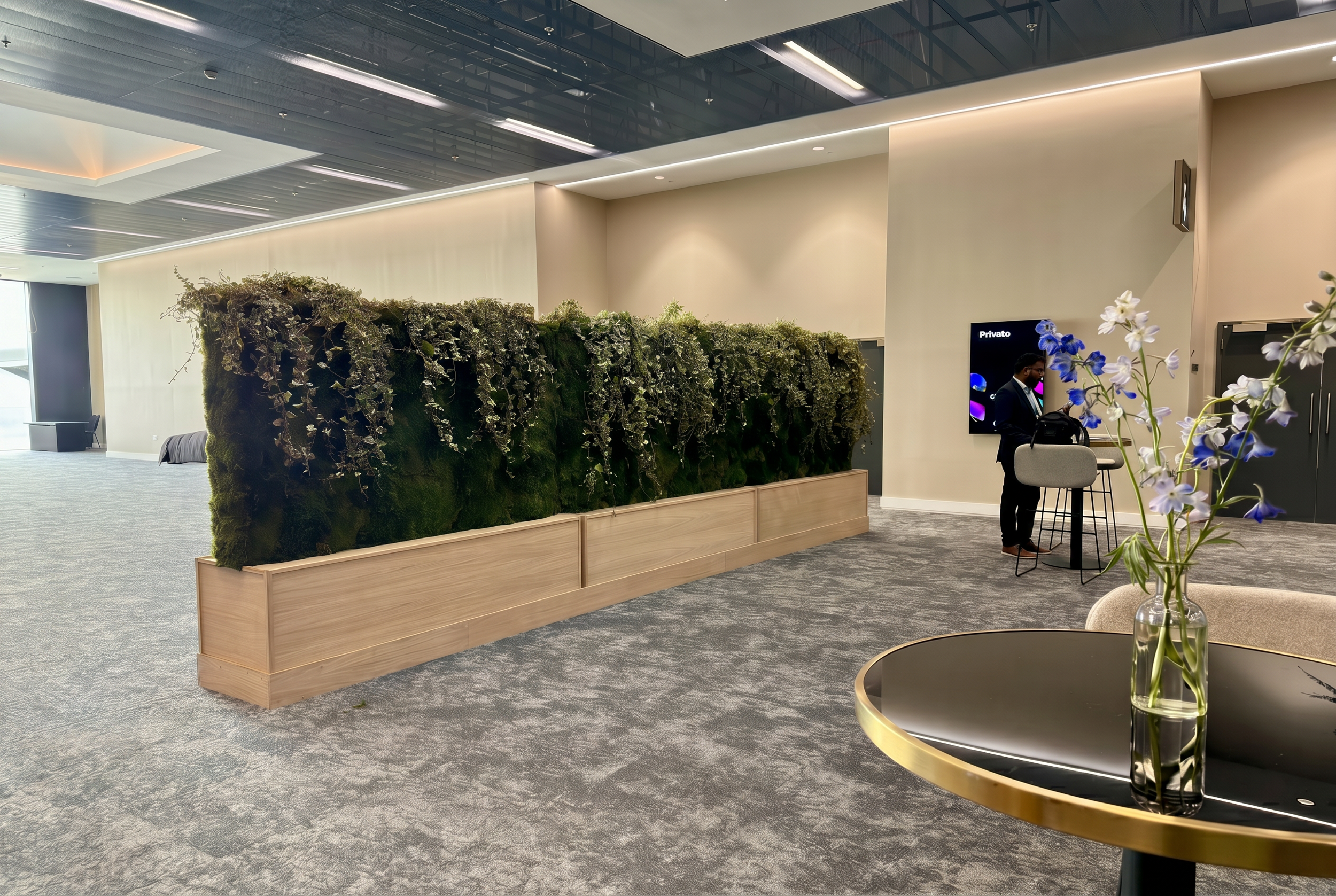 Lobby area with a large green moss and plant divider, modern ceiling with LED lighting, a man standing at a high table with barstools, and a bouquet of white and purple flowers on a round table in the foreground.