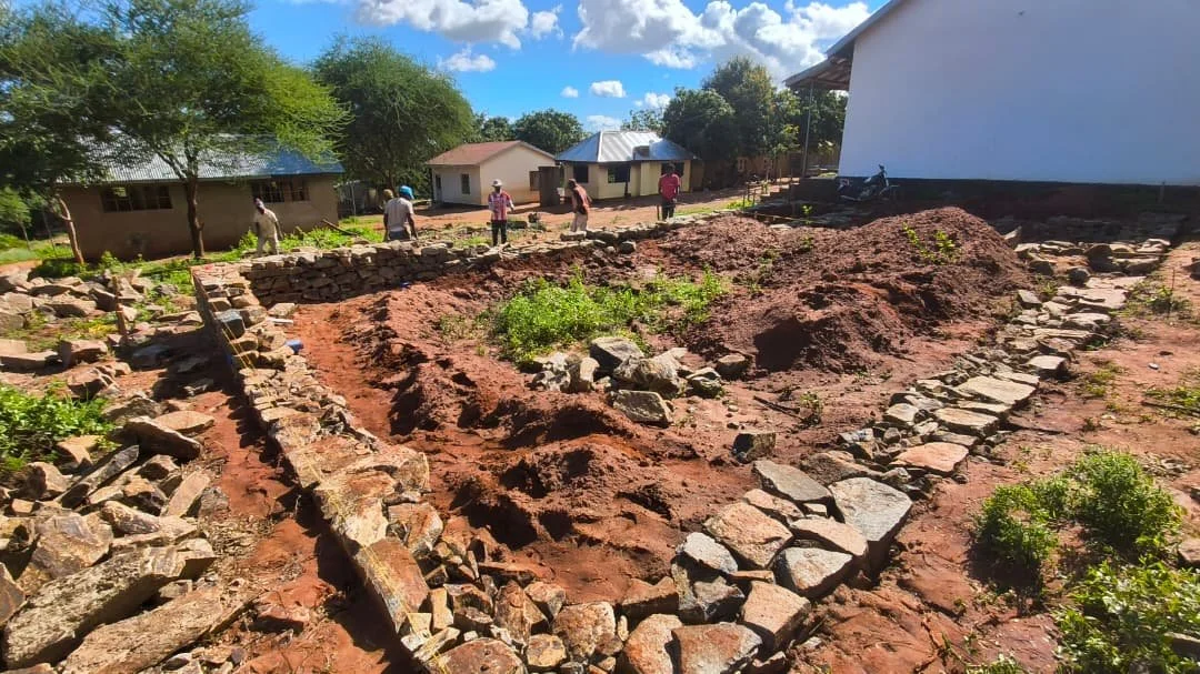 Archaeological excavation site with stone walls, tools, and workers over a dirt ground, with small trees and buildings in the background on a sunny day.