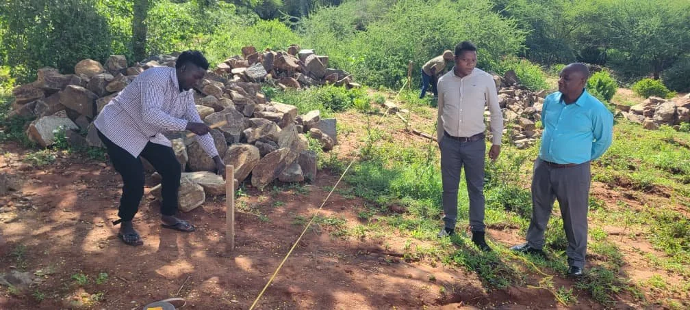 Three men outdoors on a construction site or land survey, with one man measuring and marking the land, while two others stand and observe. In the background, there are rocks and green vegetation.