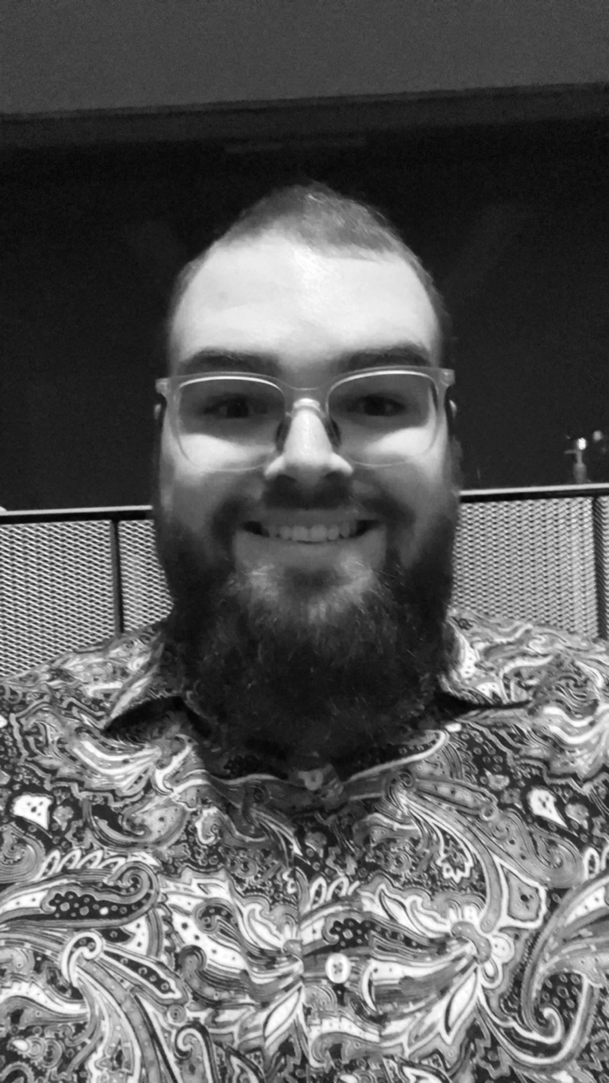 A man with glasses and a beard smiling at the camera, wearing a patterned shirt, with a blurred background.