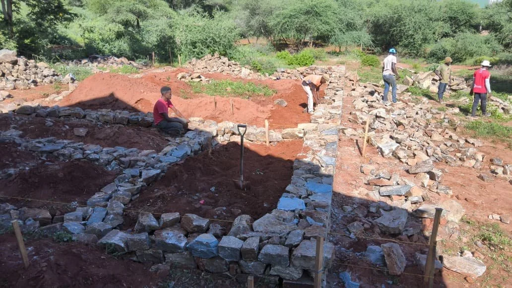 People working on a stone construction site outdoors, building foundations with rocks, surrounded by dirt and greenery.