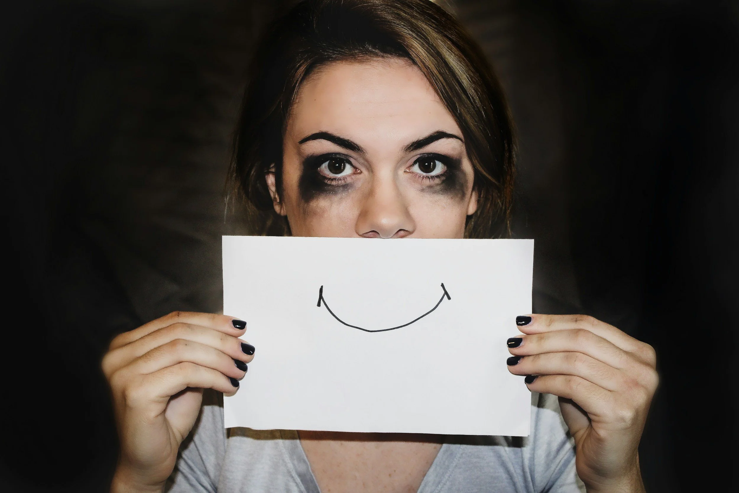 a woman with heavily smudged eye makeup holding a piece of a paper with a smile on it in front of her mouth representing healthcare worker burnout