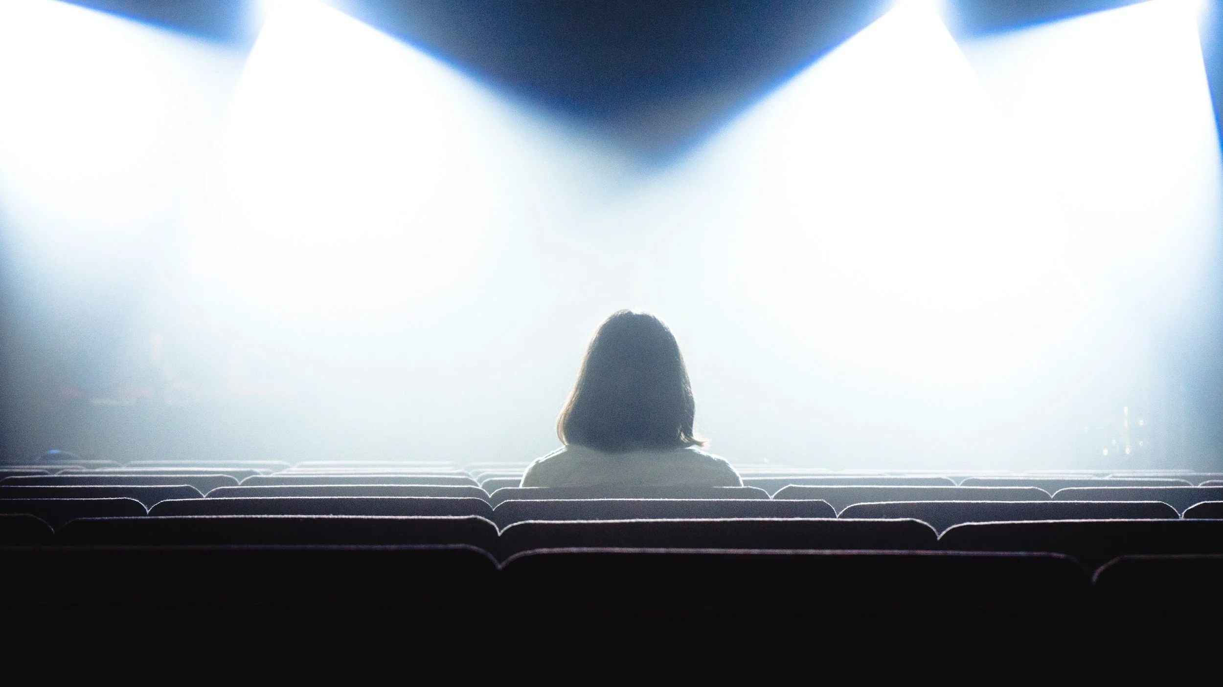 Woman sitting alone in a theater with spotlights on her, representing the feeling of being evaluated during a performance review