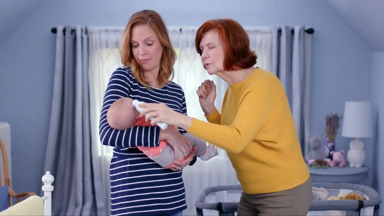 An adult appearing tense as she holds her baby and her mother corrects her, illustrating how childhood experiences of a bullying parent can shape nervous system responses well into adult life