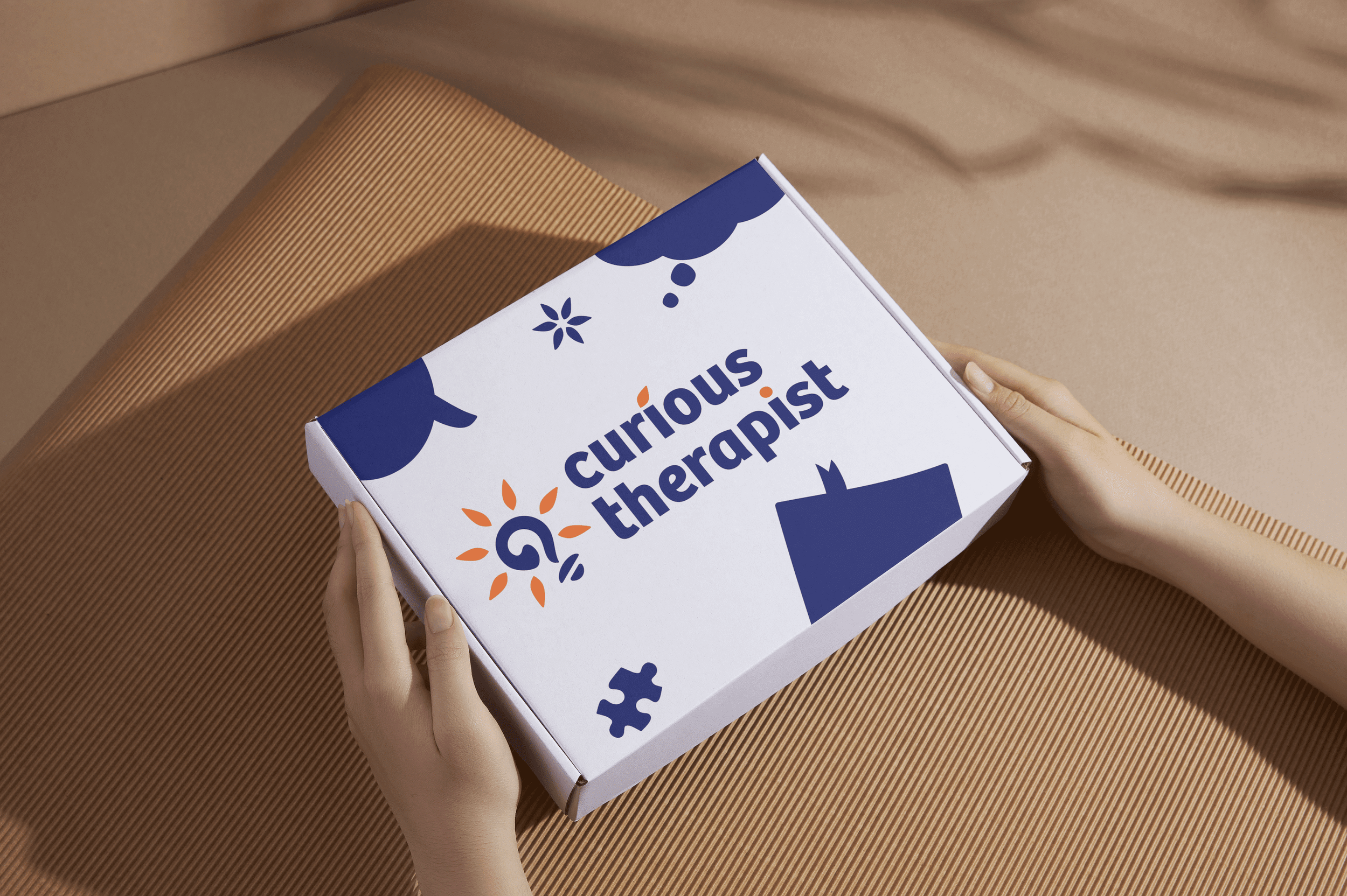 Hands holding a white box with the words 'curious therapist' in bold blue and orange letters, featuring graphic elements such as puzzle pieces, a light bulb, and clouds, on a beige textured surface.