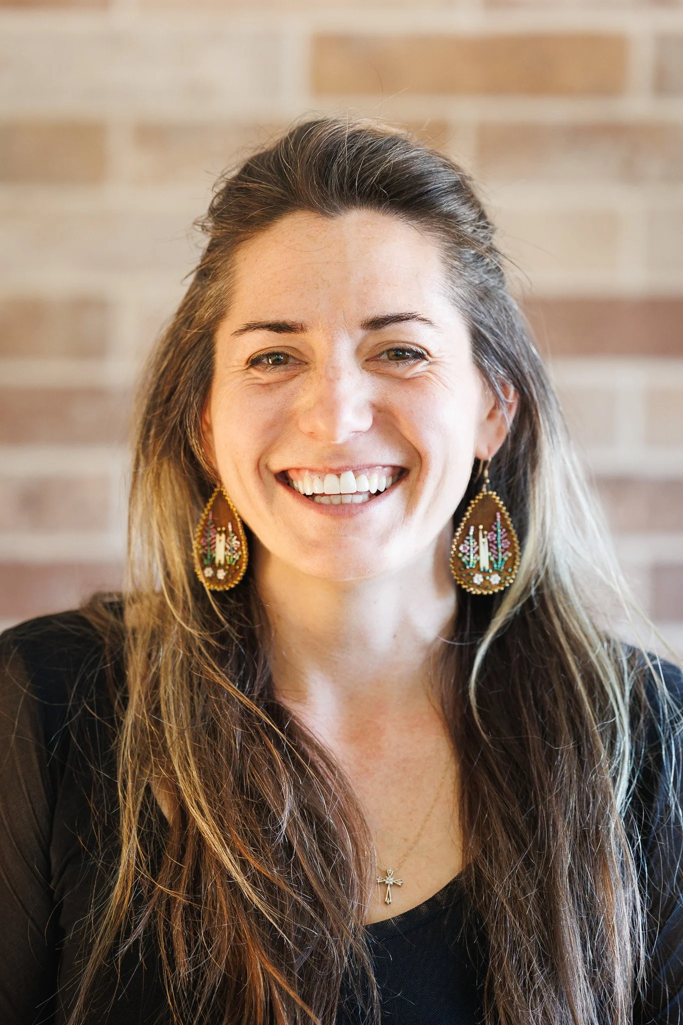A woman with long brown hair, smiling outdoors with blurry trees and sky in background, wearing a striped jacket and white top.
