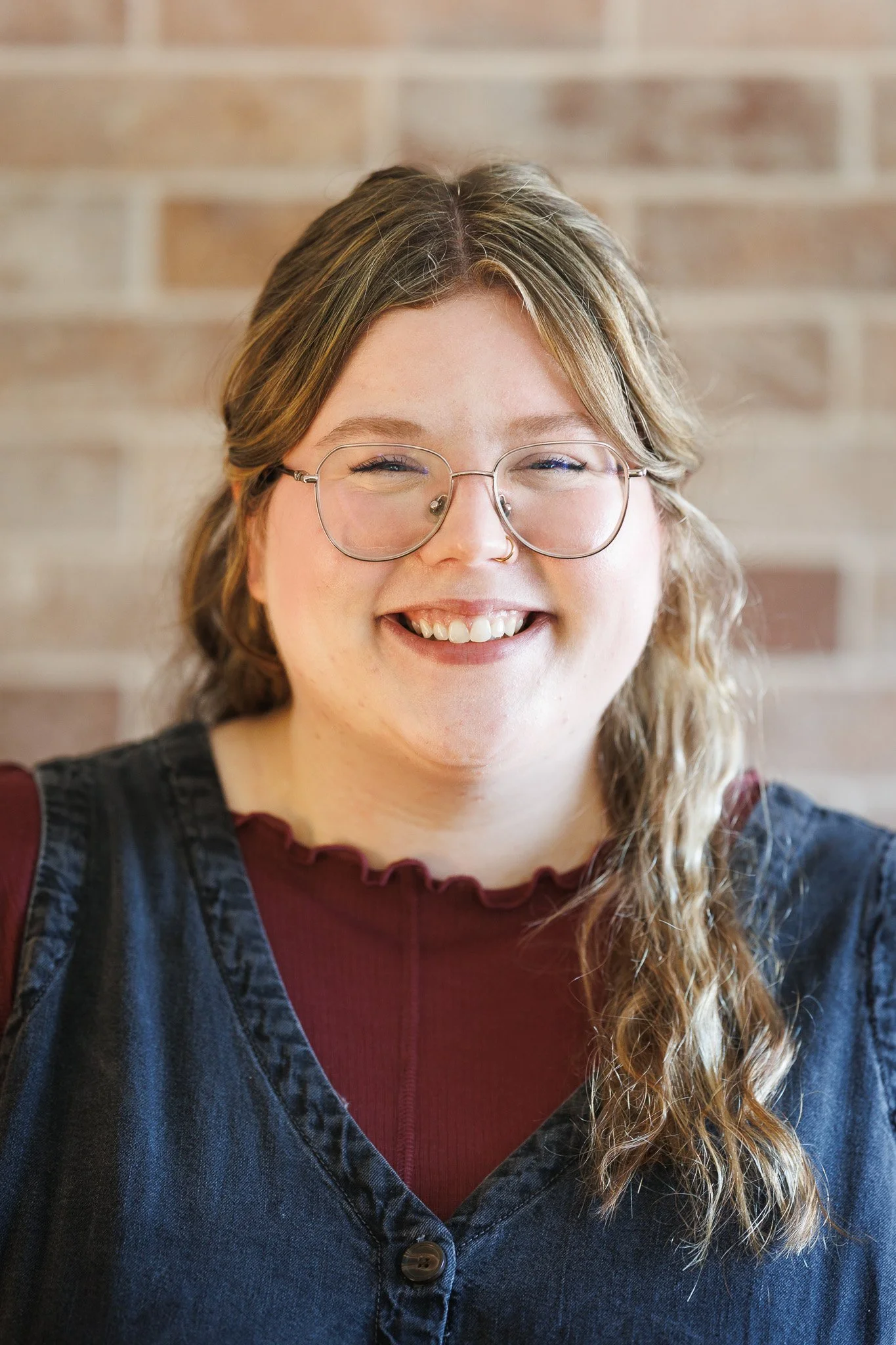 Smiling young woman with glasses and long wavy brown hair outdoors in fall or winter, wearing a plaid jacket over a black top.