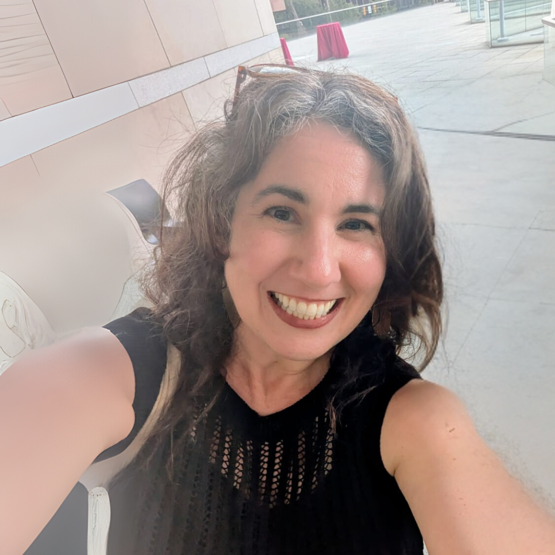 Una mujer con cabello rizado y oscuro, sonriendo, lleva puesta una blusa negra sin mangas, en un lugar con paredes de color beige y ventanas grandes al fondo, durante el día.