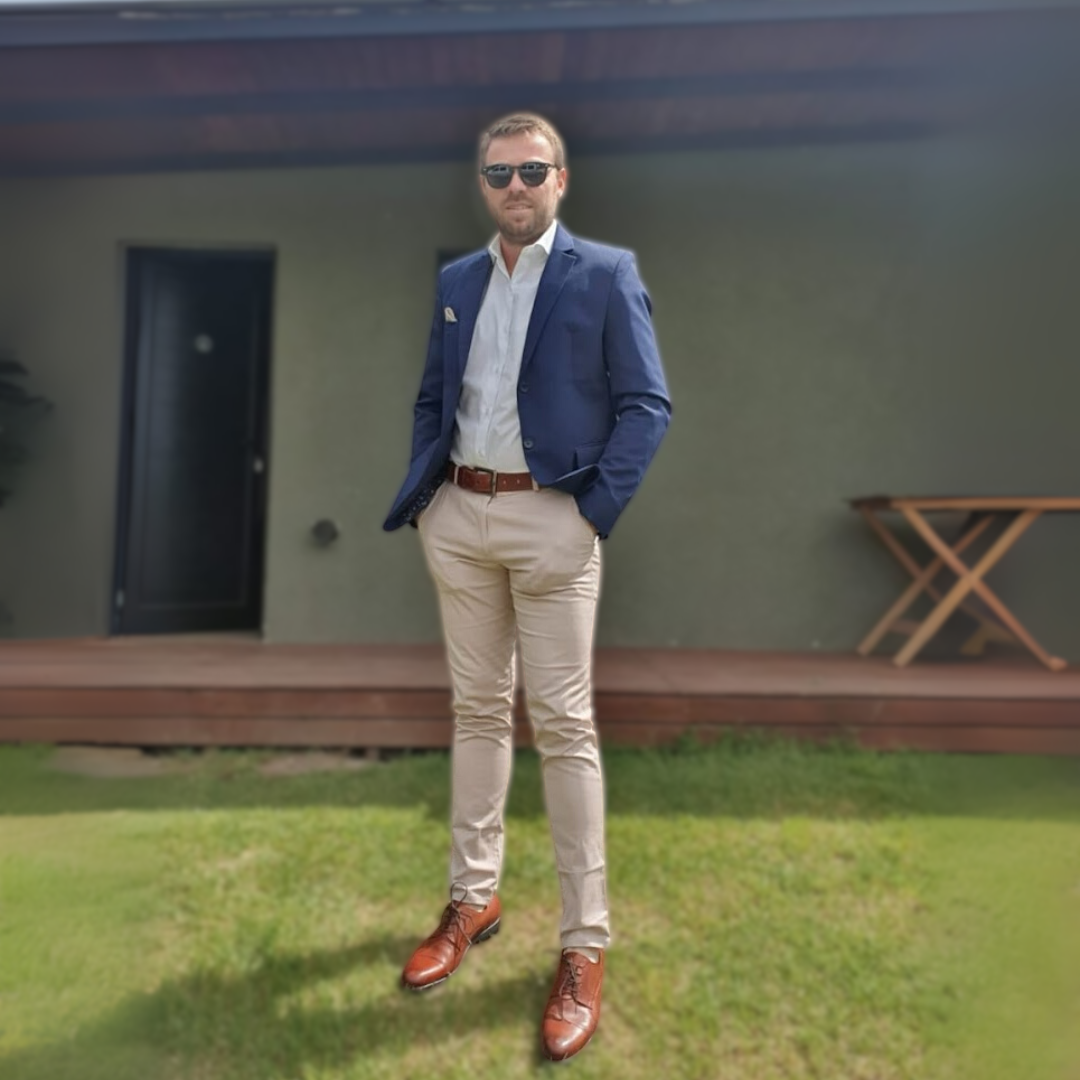 Hombre vestido con saco azul, camisa blanca, pantalones beige y zapatos marrones, con gafas de sol, posando en un patio con césped y fondo de pared gris.
