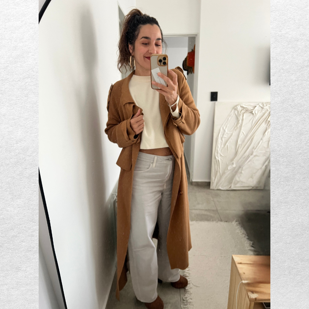 Mujer con cabello rizado con una coleta alta, vestida con blusa blanca, pantalones beige y abrigo marrón, tomándose una selfie frente a un espejo en una habitación moderna.