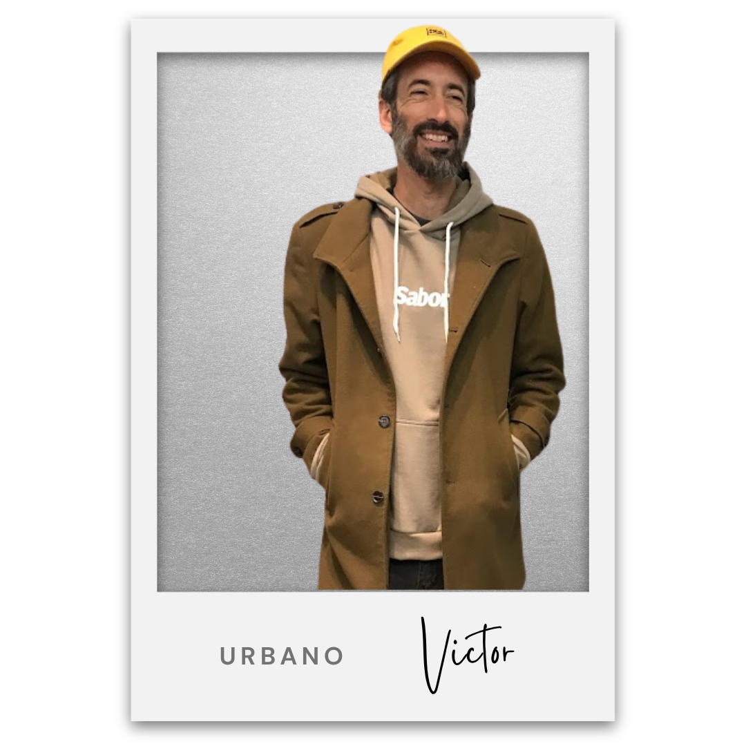 Hombre sonriendo con barba, llevando una gorra amarilla, sudadera beige con capucha y abrigo marrón, en un fondo gris. Victor Mochkofsky
