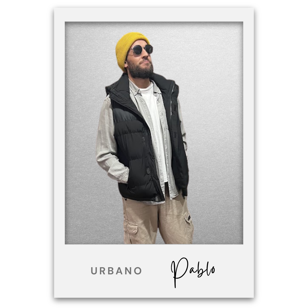 Un hombre de vestimenta urbana con gafas de sol, gorro amarillo y barba, vistiendo una chaqueta negra, camisa gris y pantalones beige, posa con las manos en los bolsillos, fondo gris, en un marco que dice 'Urbano Pablo'. Pablo Bocco