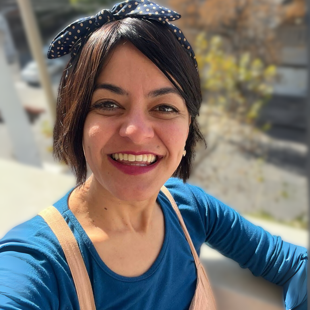 Mujer sonriendo posando al aire libre con fondo de árboles y paredes de piedra, lleva bandana con puntos y camiseta azul.