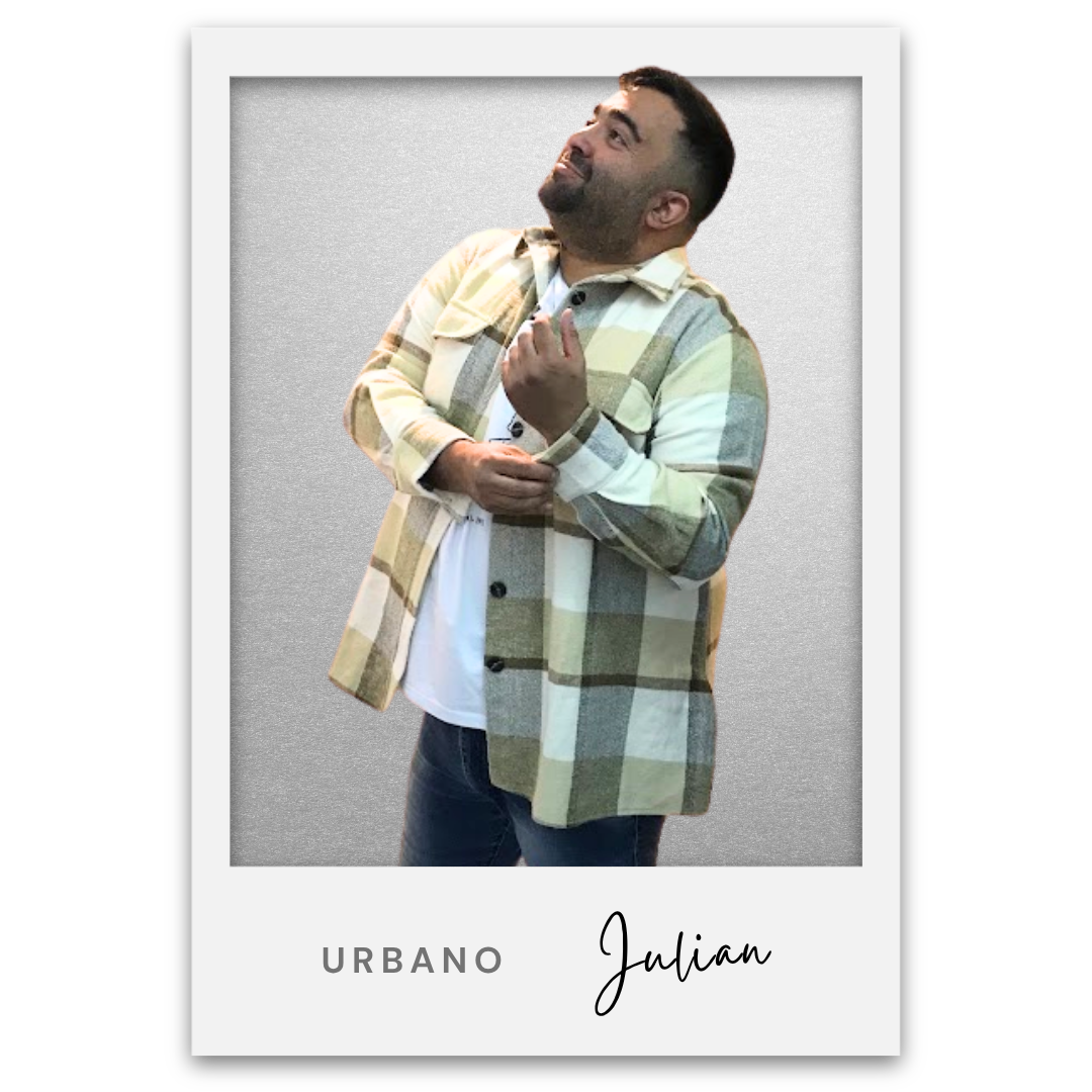 Hombre sonriente con barba, usando una chamarra a cuadros en tonos beige, gris y verde, con una camiseta blanca debajo, de pie contra un fondo gris. Texto en la parte inferior dice 'URBANO Julian'. Julian Bronstein