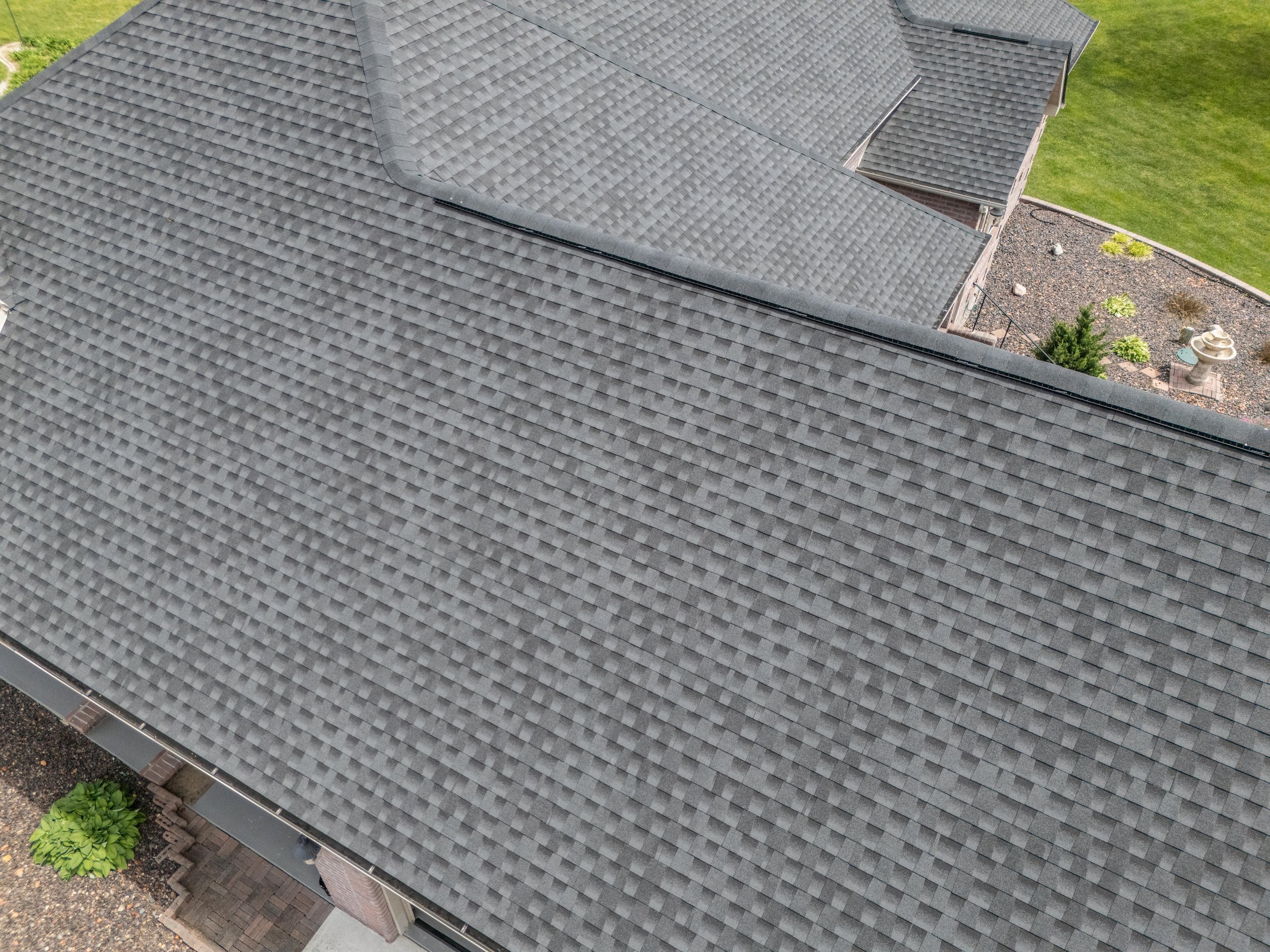 Top view of a residential roof with gray asphalt shingles, surrounded by a landscaped yard with green grass and a stone garden feature.